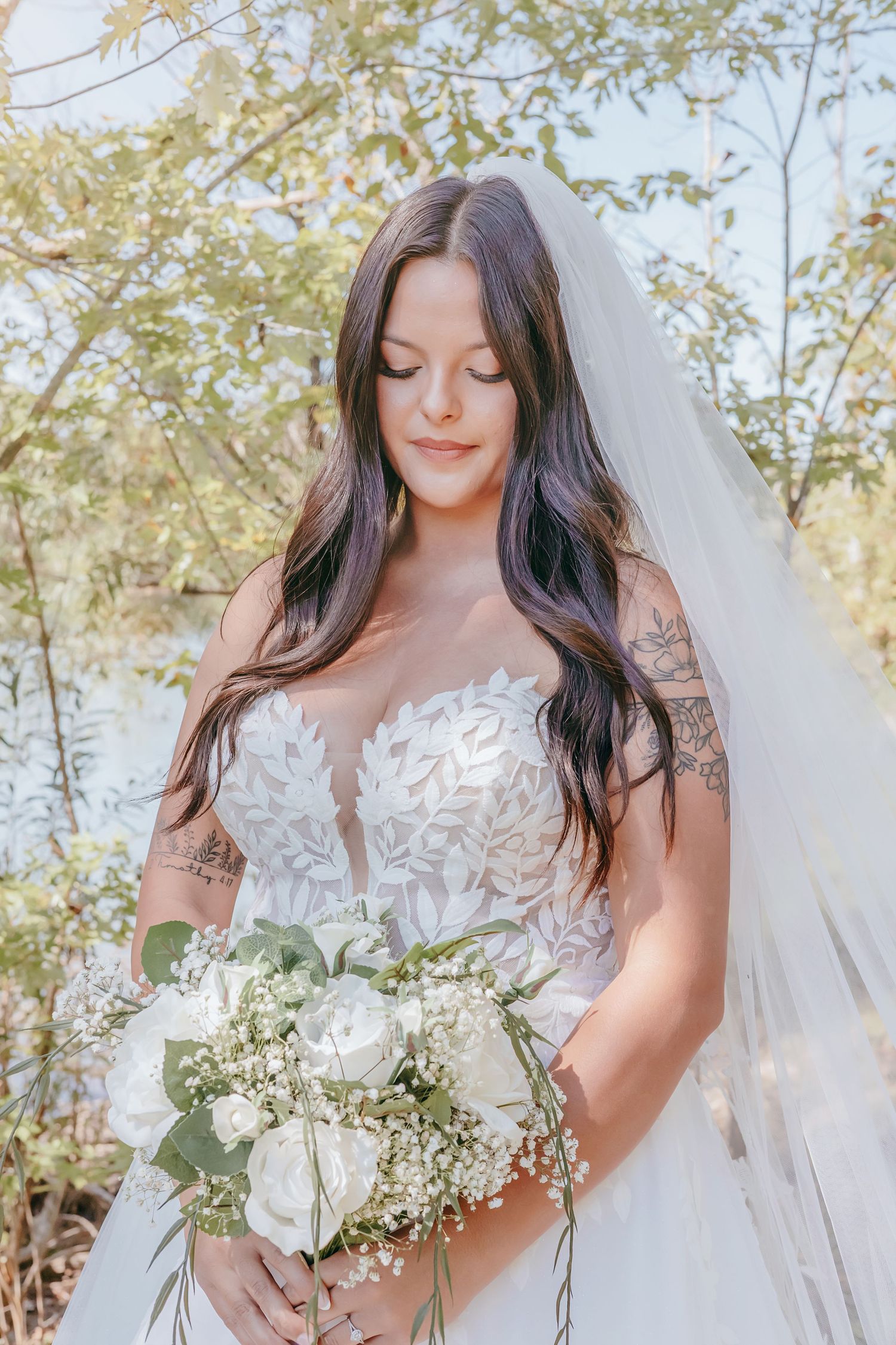 Bride with floral tattoos holding white rose bouquet in lace gown and veil near sunlit trees.