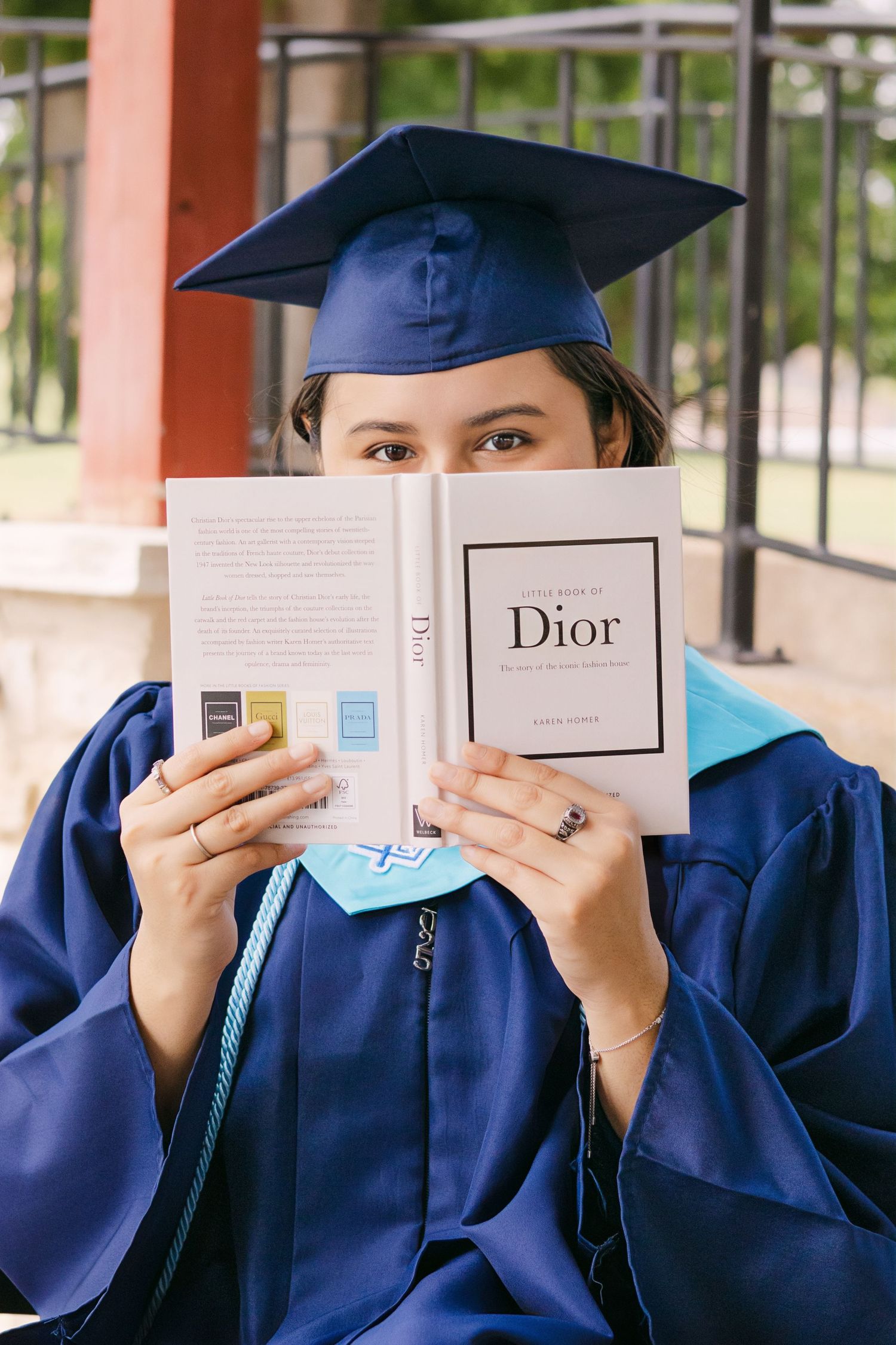 Graduate in blue cap and gown holds a Dior book covering her face outdoors.