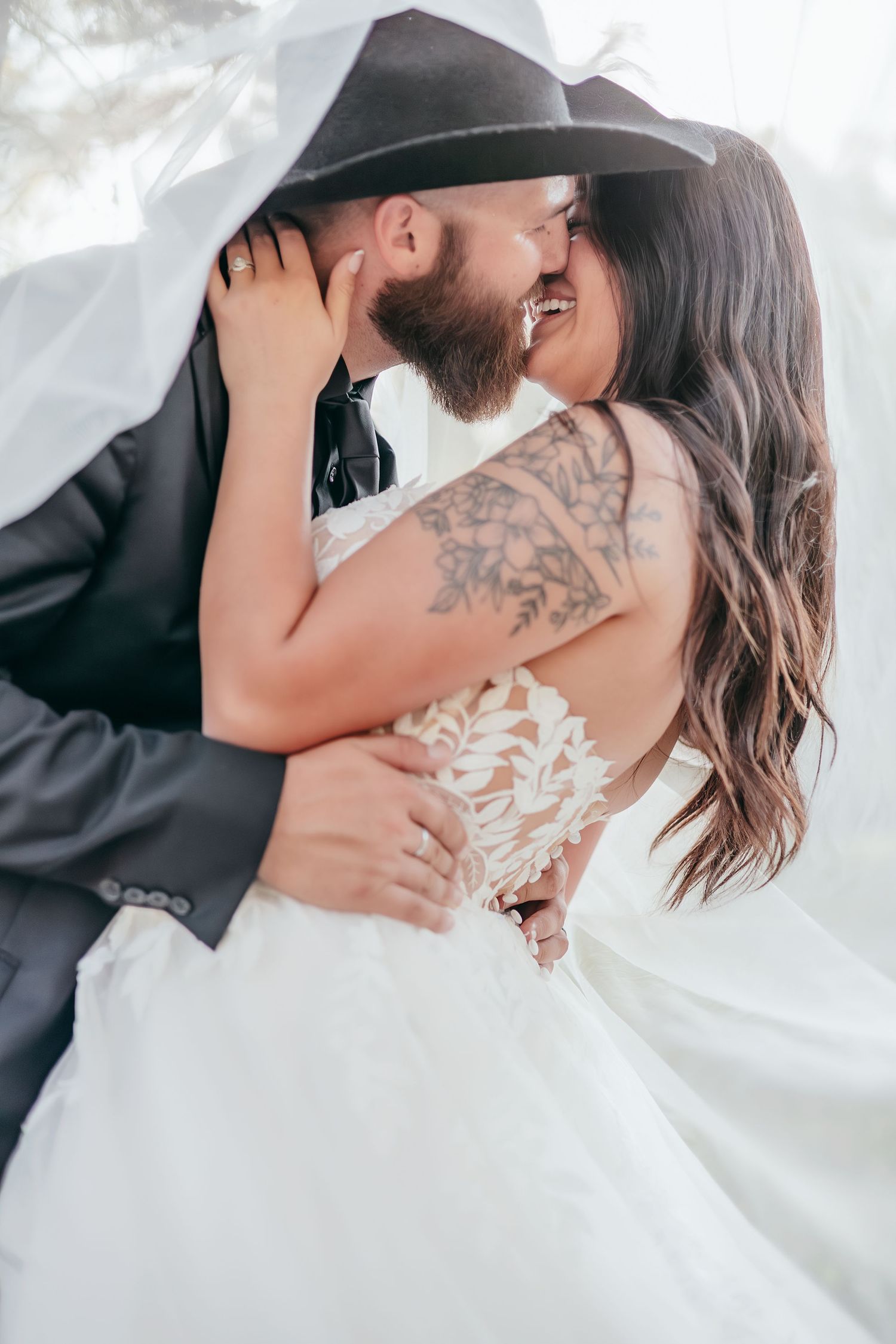 Bride with floral arm tattoo and groom in black cowboy hat embracing and kissing under wedding veil.