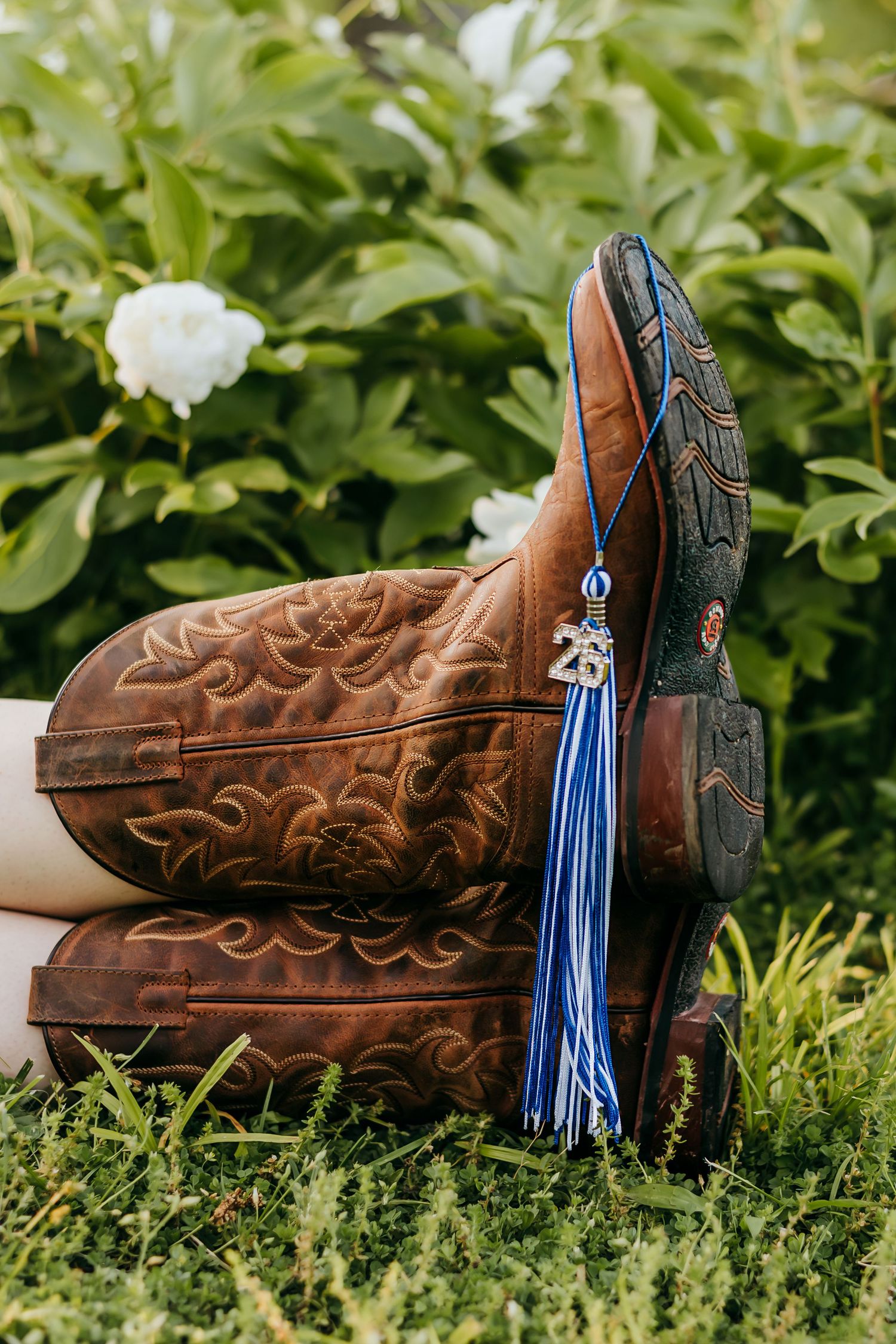 Brown leather cowboy boots with blue graduation tassel resting on green grass near white flowers.
