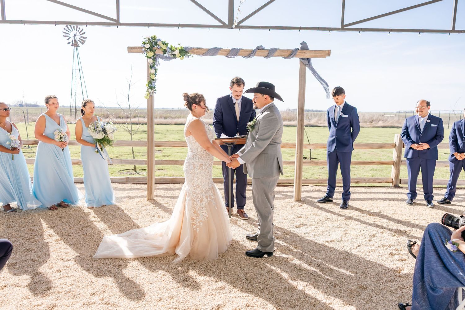 Bride and groom exchange vows under a rustic wooden arch with bridesmaids in dusty blue dresses.