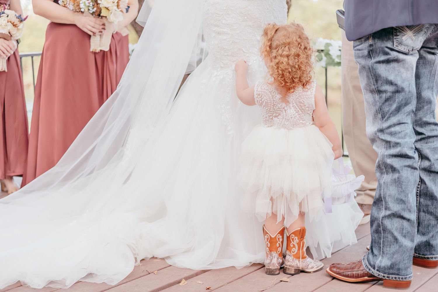 Flower girl in white tulle dress and cowboy boots stands beside bride's lace wedding gown train.