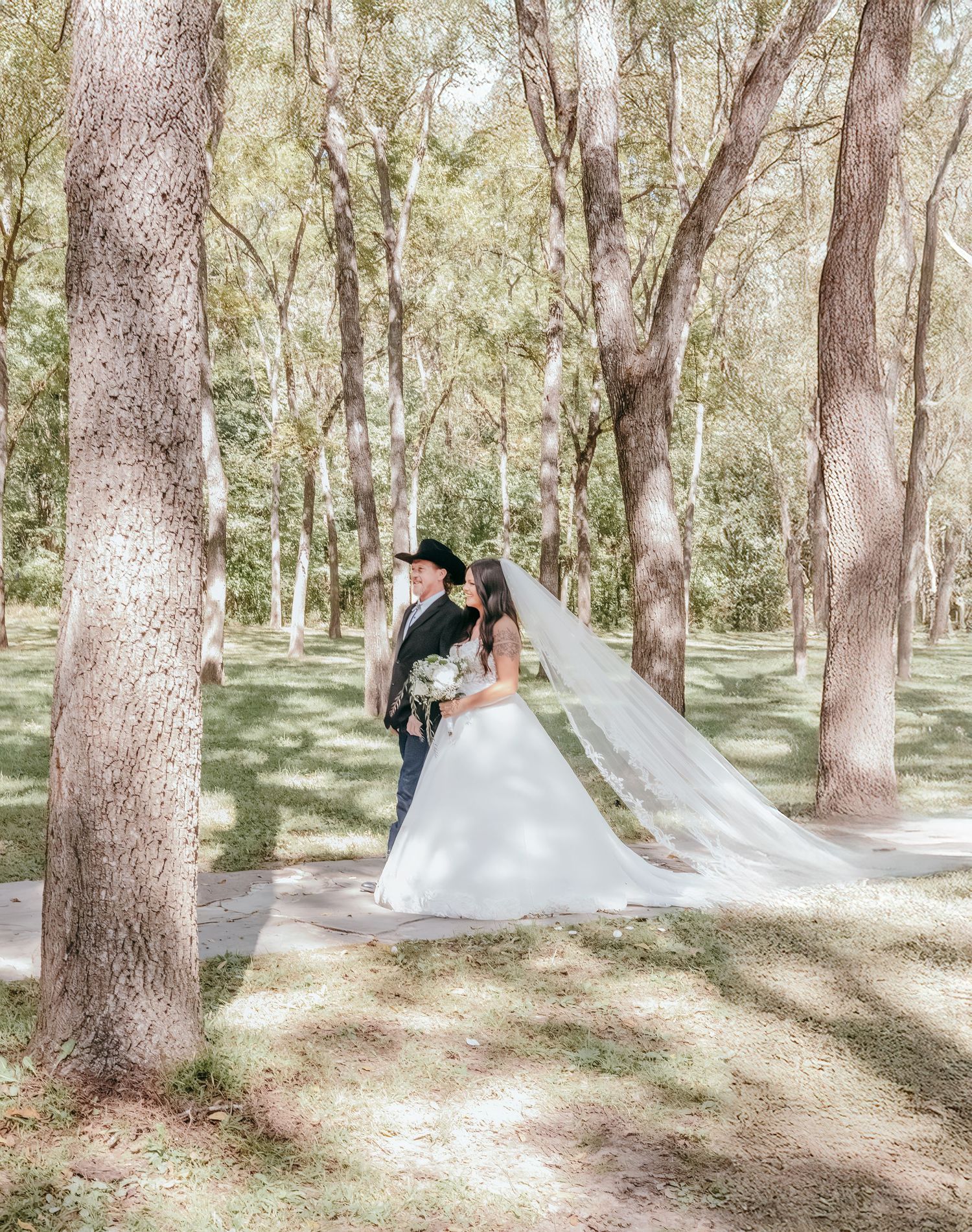 Bride in flowing white gown and veil stands with groom in black cowboy hat among tall trees.