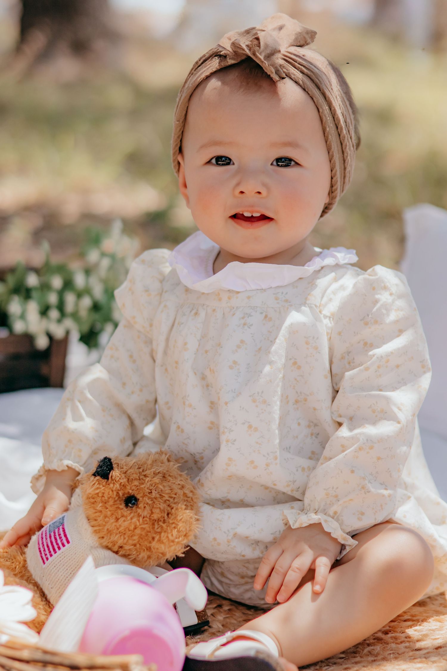 Adorable baby girl in floral dress with bow headband sitting outdoors holding a teddy bear.