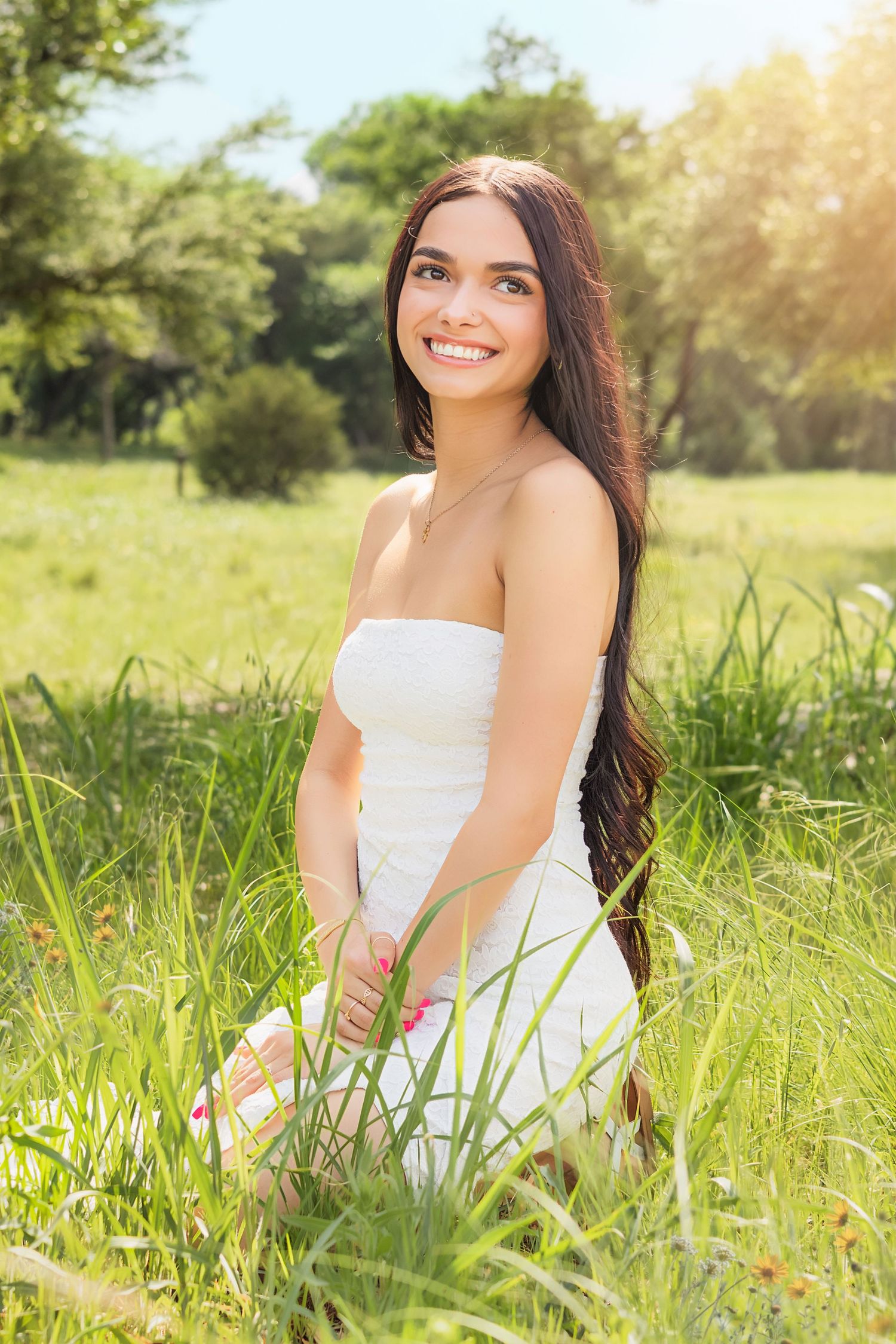 Young woman in white strapless dress sitting in lush green grass field, smiling outdoors on a sunny day.
