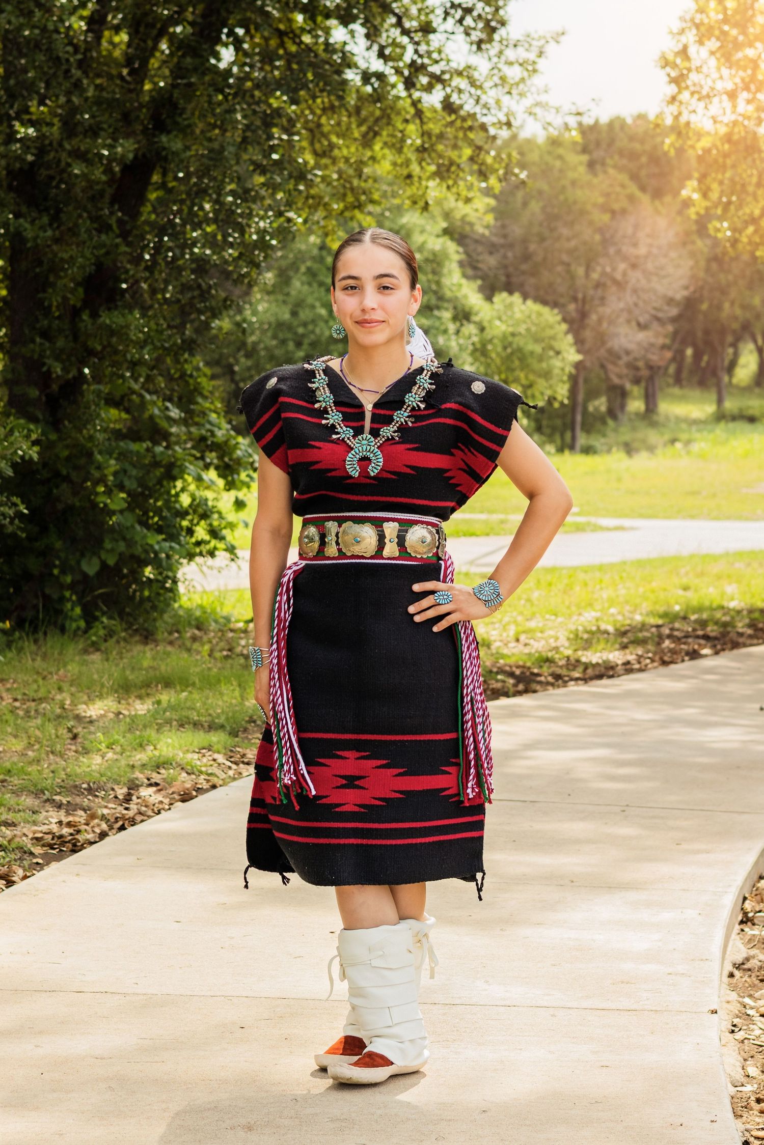 Young woman in traditional Native American dress with turquoise jewelry and moccasins standing on a pathway outdoors.