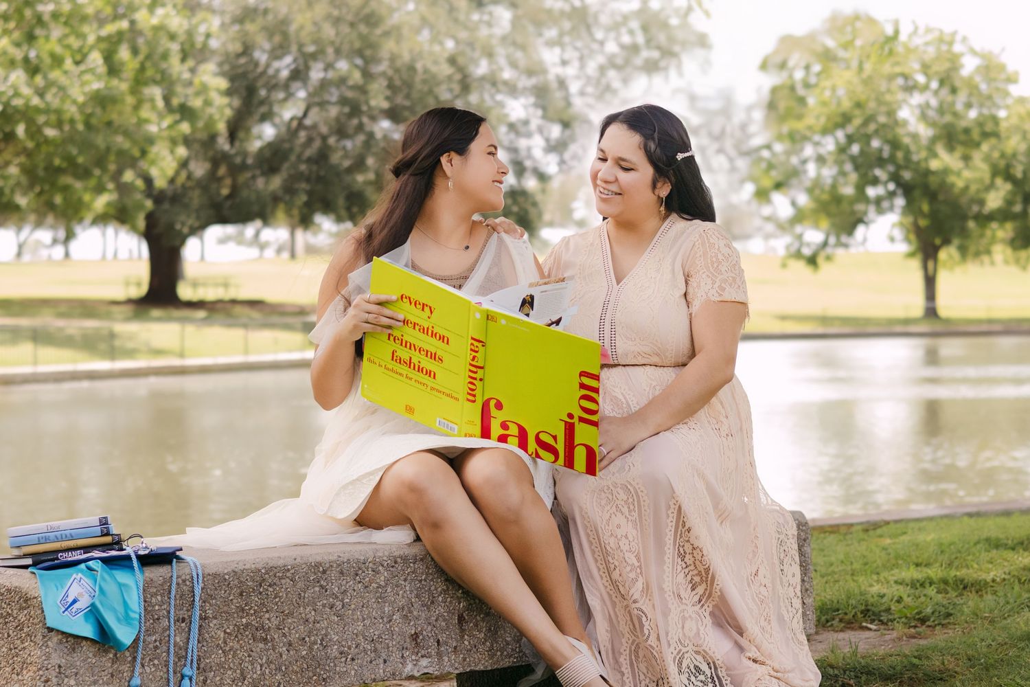 Two young women sit on stone bench sharing a colorful book beside a park pond.
