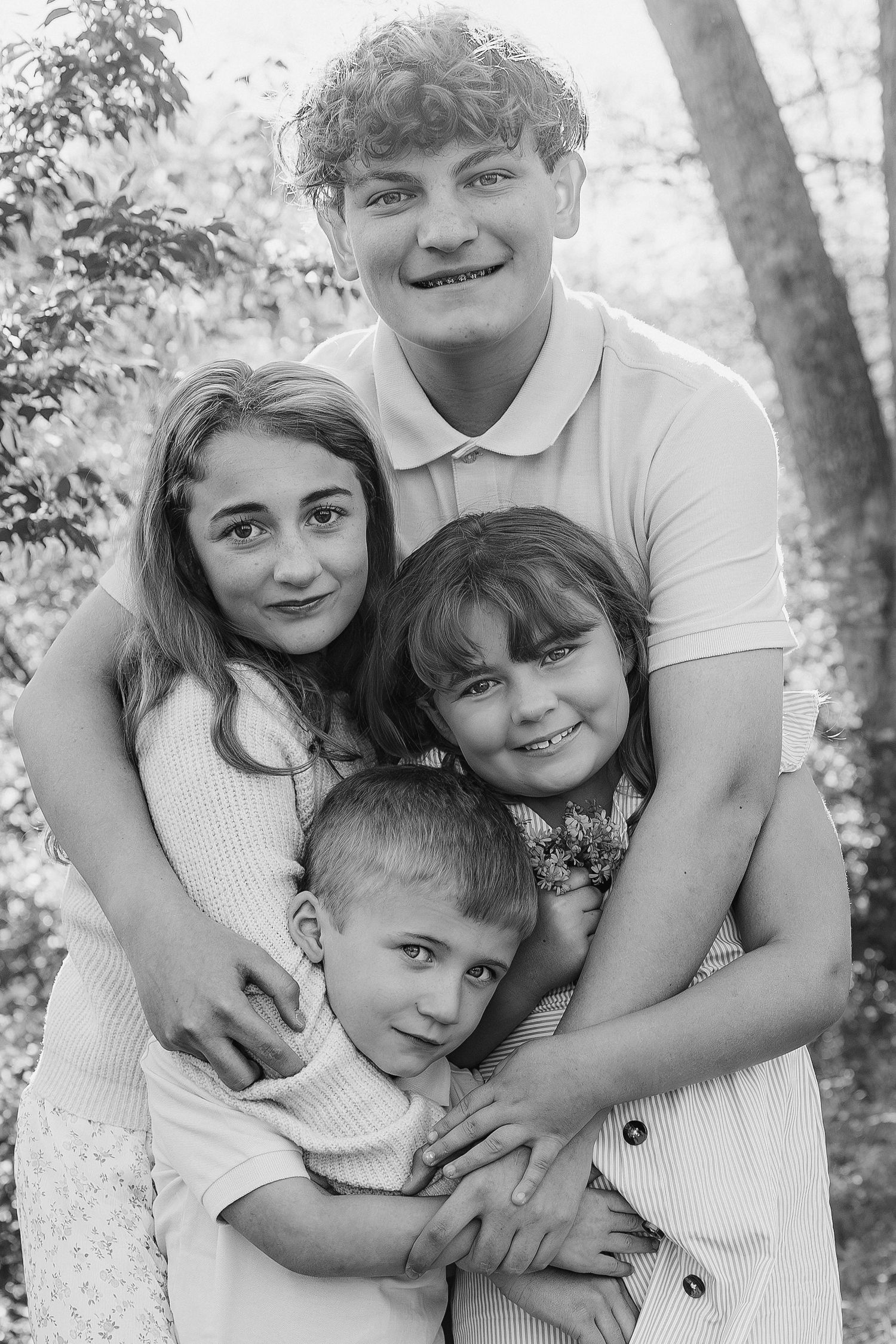 Black and white family portrait of four siblings hugging outdoors among trees, capturing a warm and joyful moment.