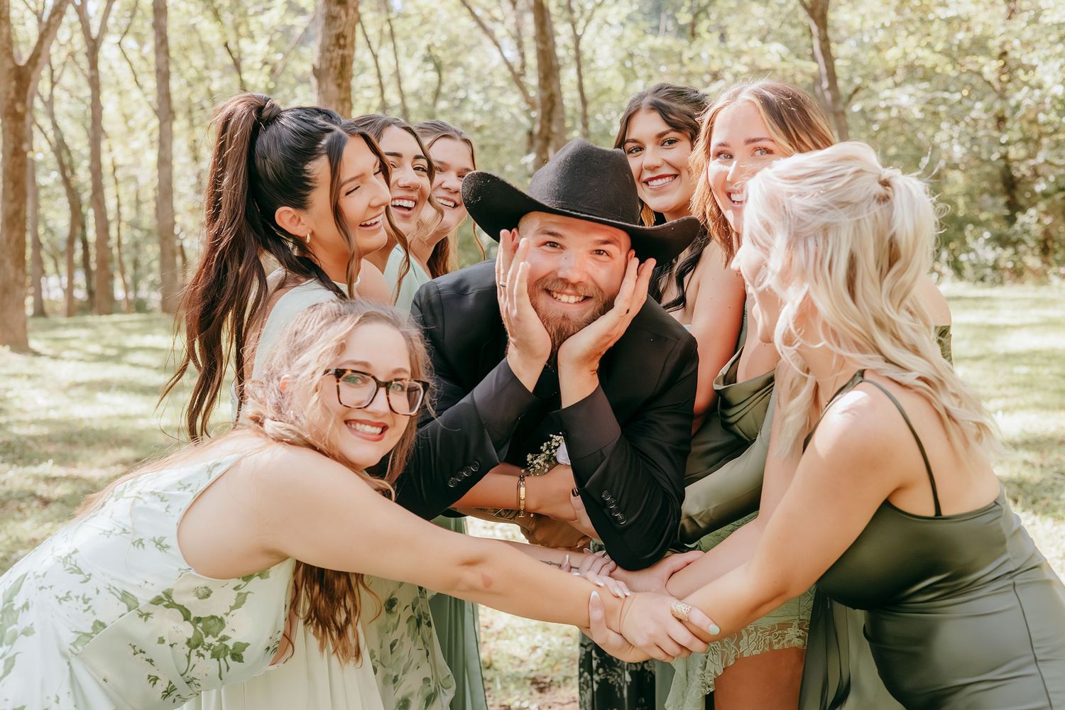 Bearded groom in black cowboy hat surrounded by laughing bridesmaids in sage green dresses in wooded setting.