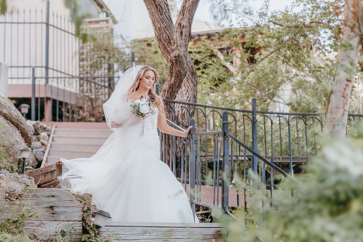 Smiling bride in white mermaid gown holding bouquet on wooden steps near trees.