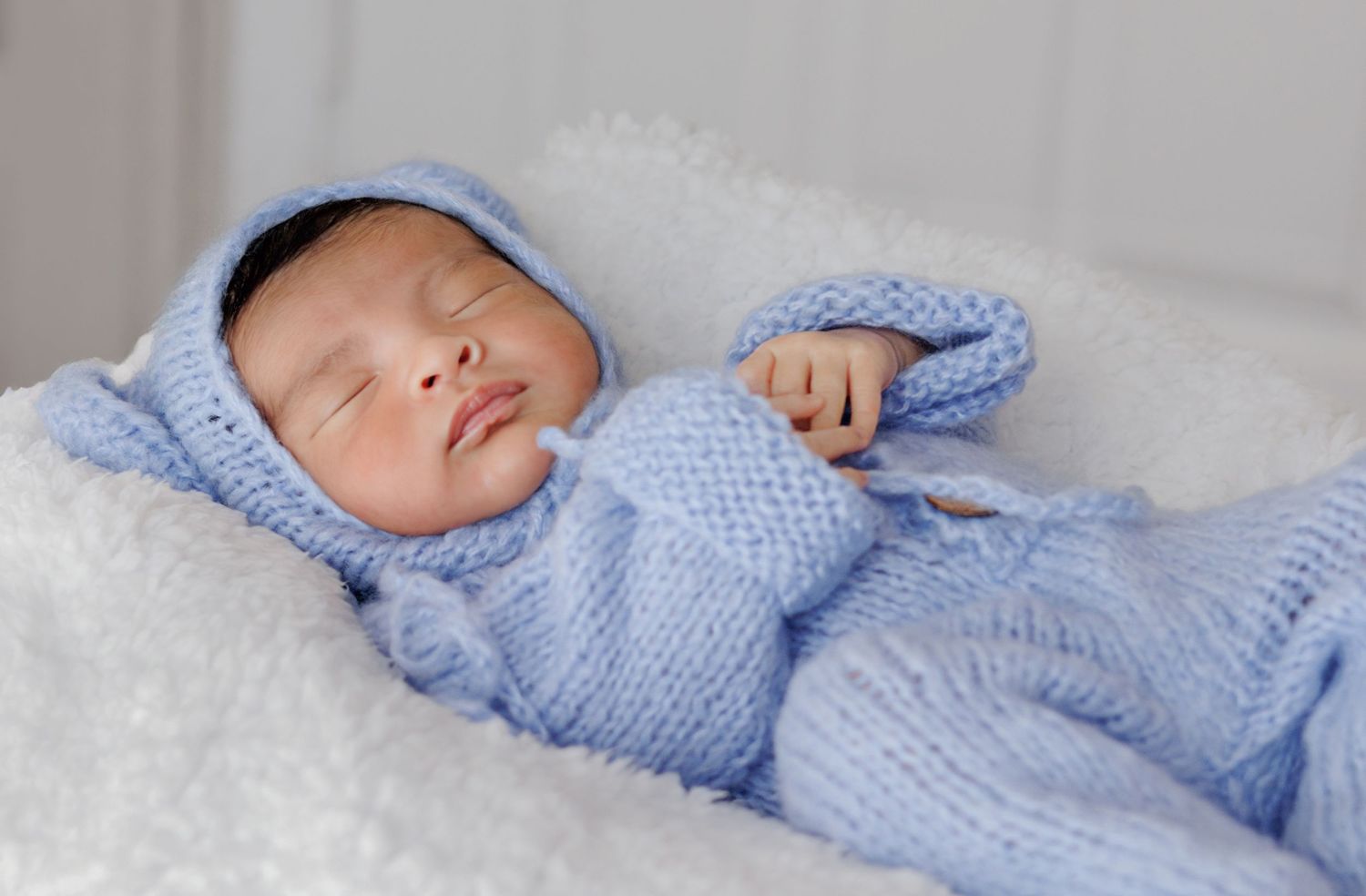 Newborn baby sleeping peacefully in a blue knit outfit and hat on a soft white blanket.