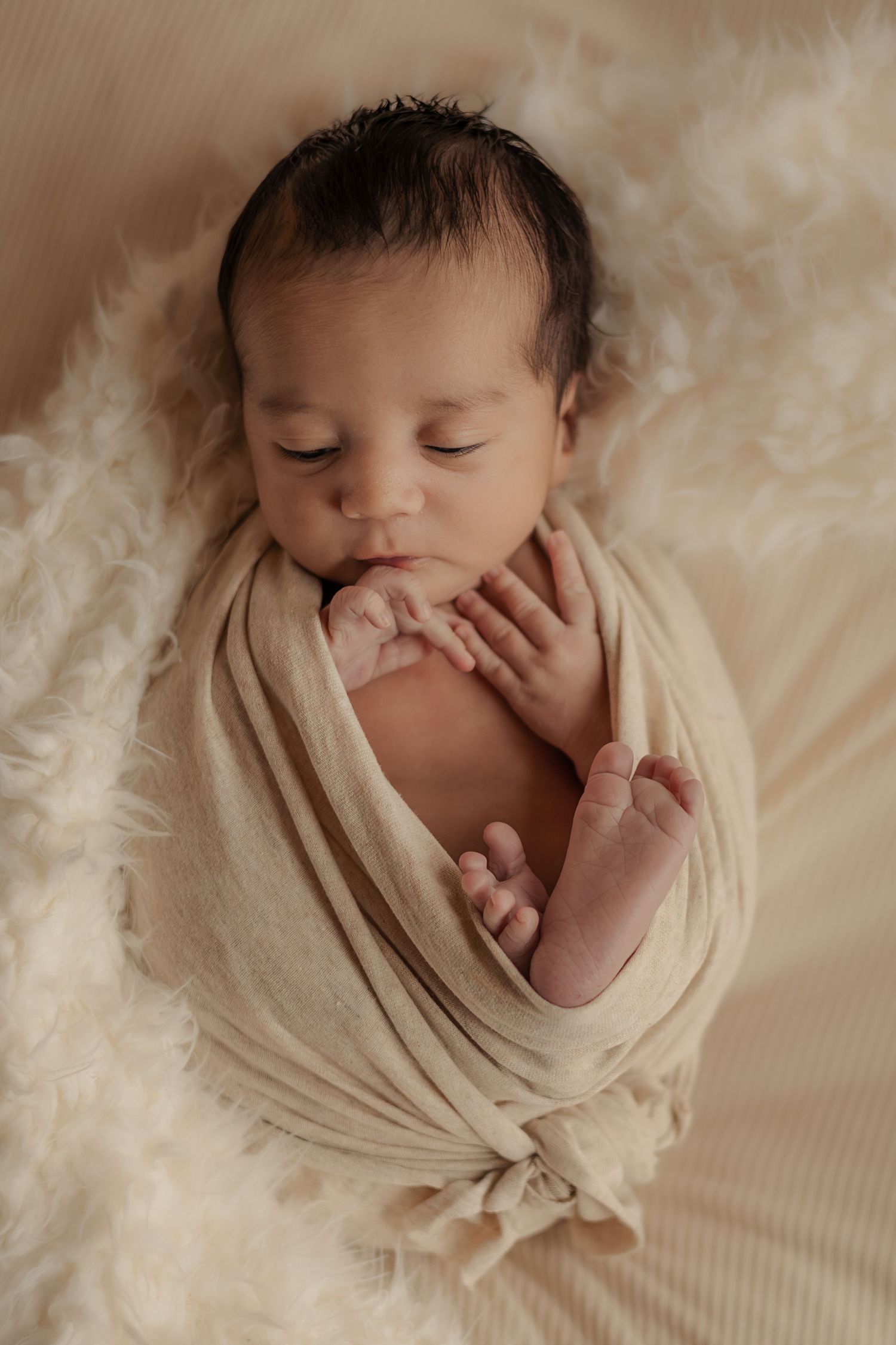 Sleeping newborn baby swaddled in a cream wrap with tiny hands visible on a soft fluffy blanket.