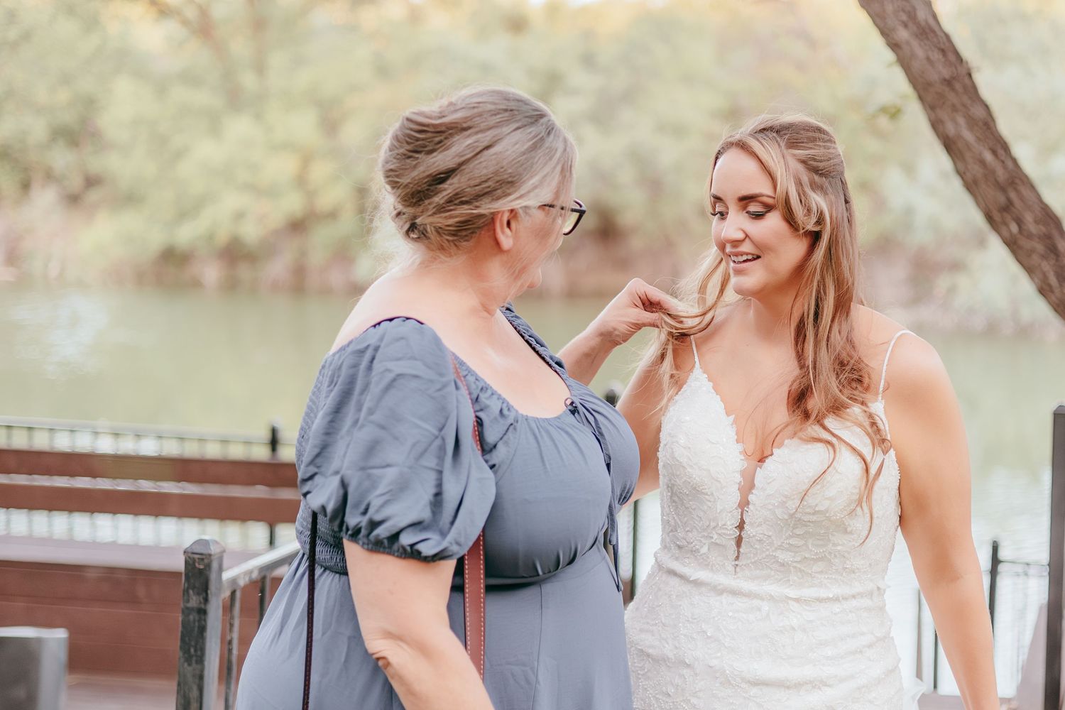A bride in a white lace gown shares a tender moment with a woman in a blue dress near a scenic waterway.