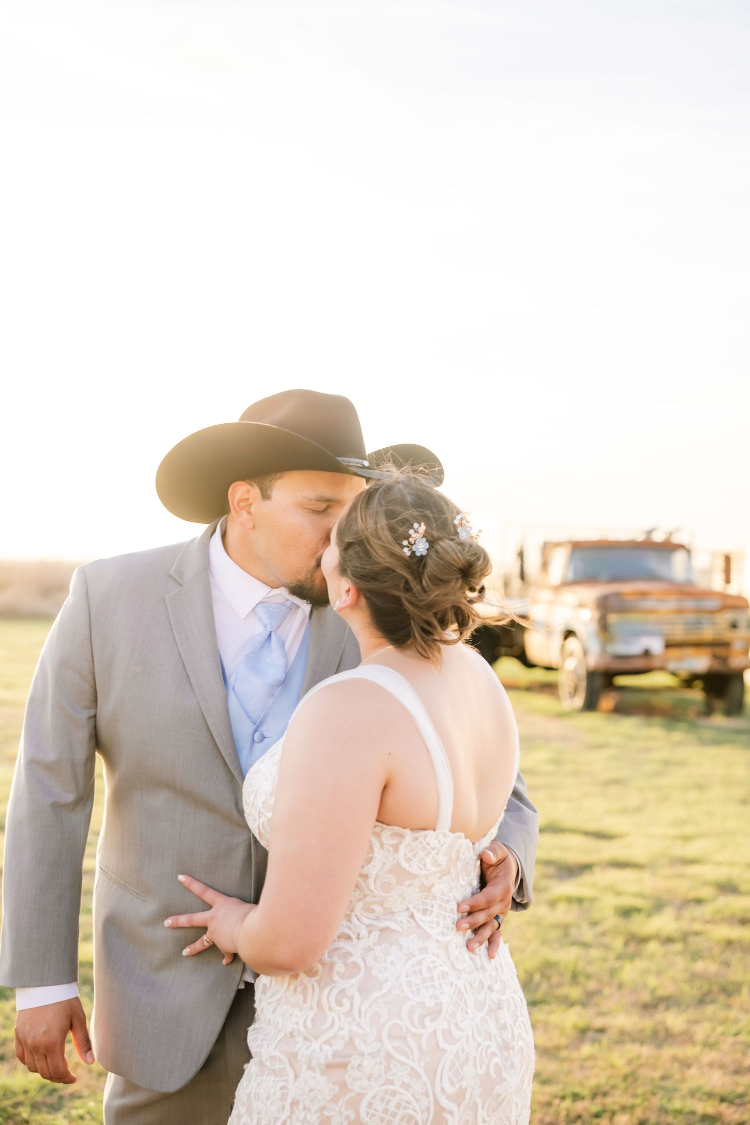 Groom in cowboy hat and gray suit kisses bride in lace gown near a vintage rusty truck at sunset.