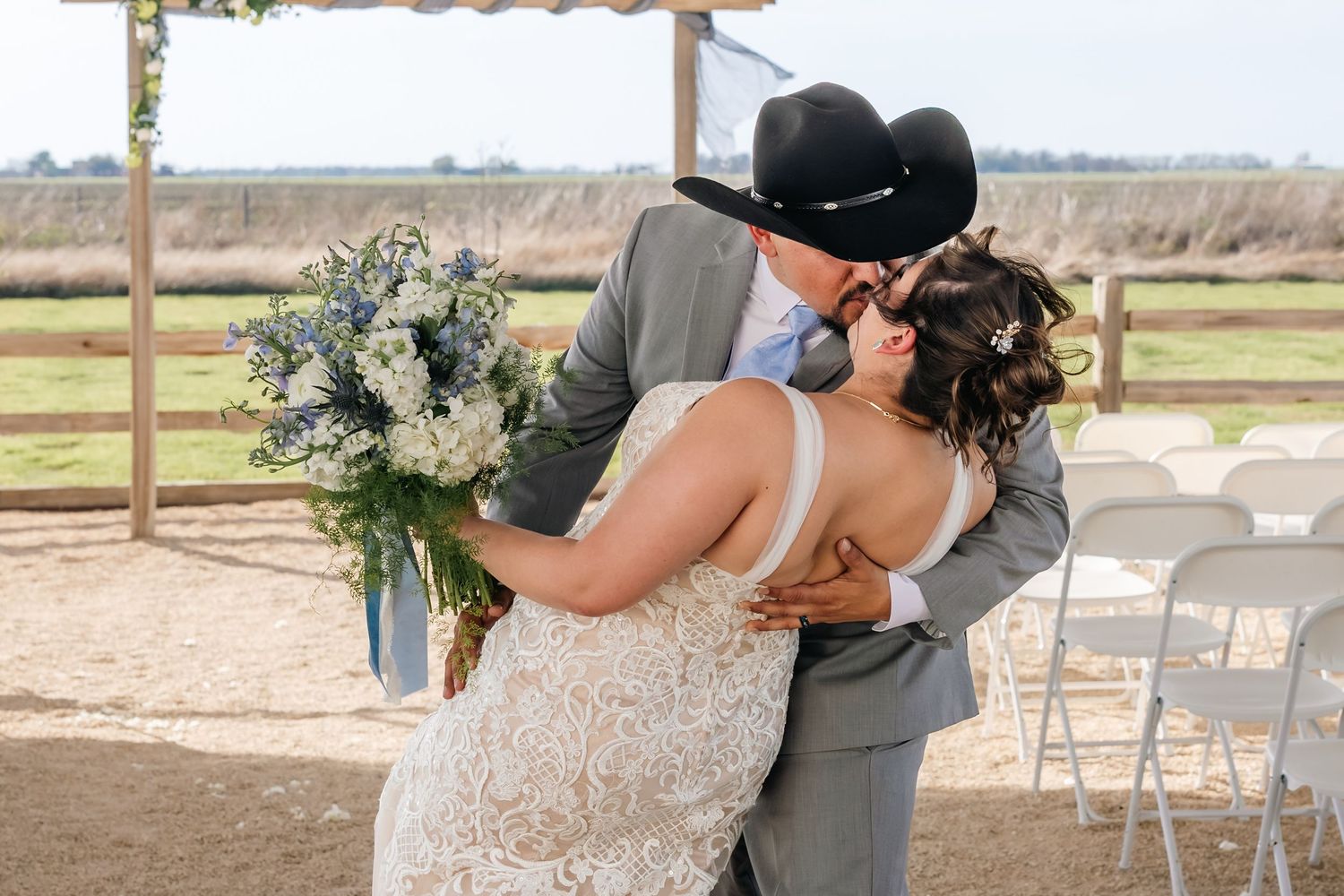 Bride and groom share first kiss at rustic outdoor wedding ceremony, groom wearing black cowboy hat and gray suit.