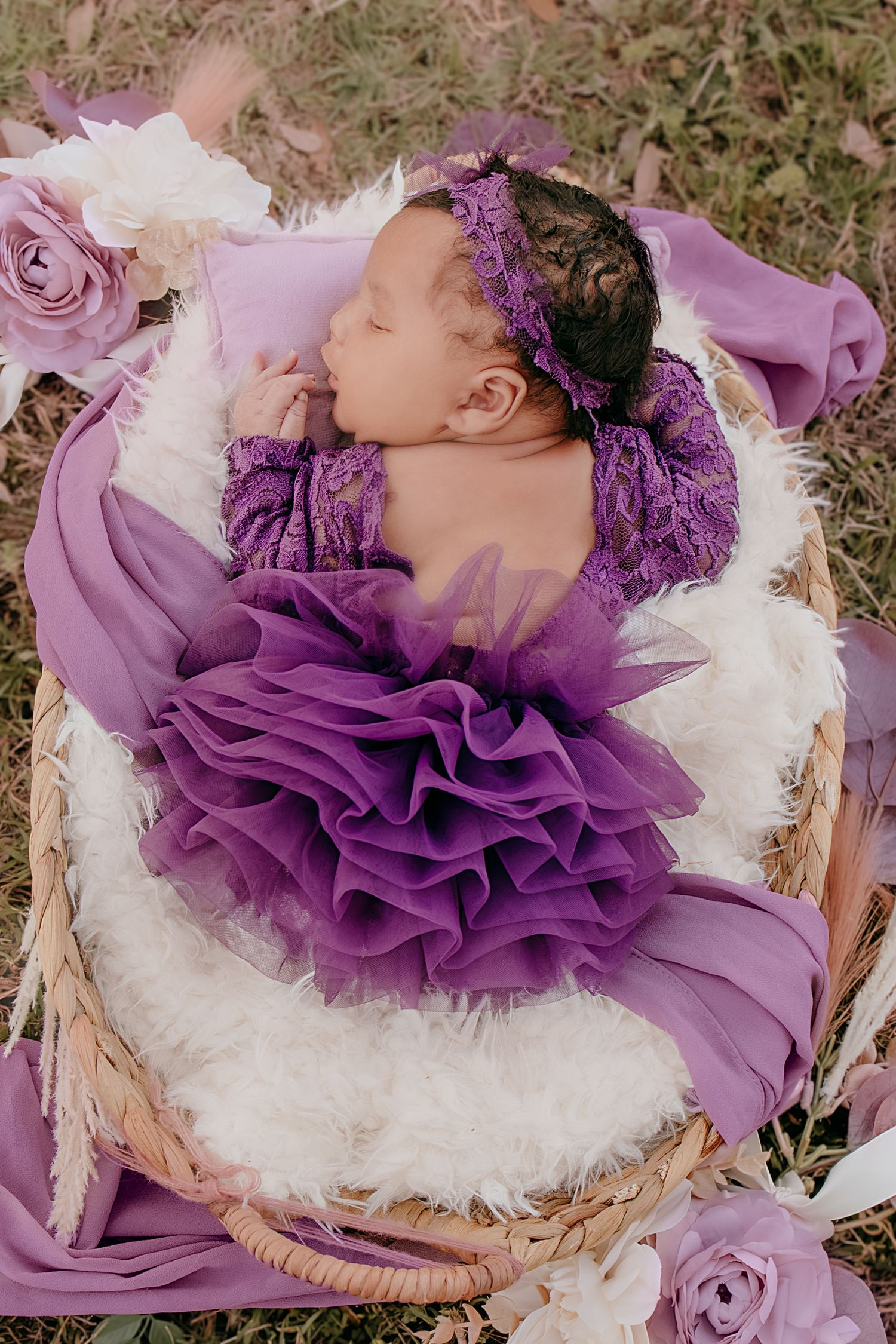 Newborn baby in purple ruffled dress sleeping in wicker basket surrounded by pink and white flowers.