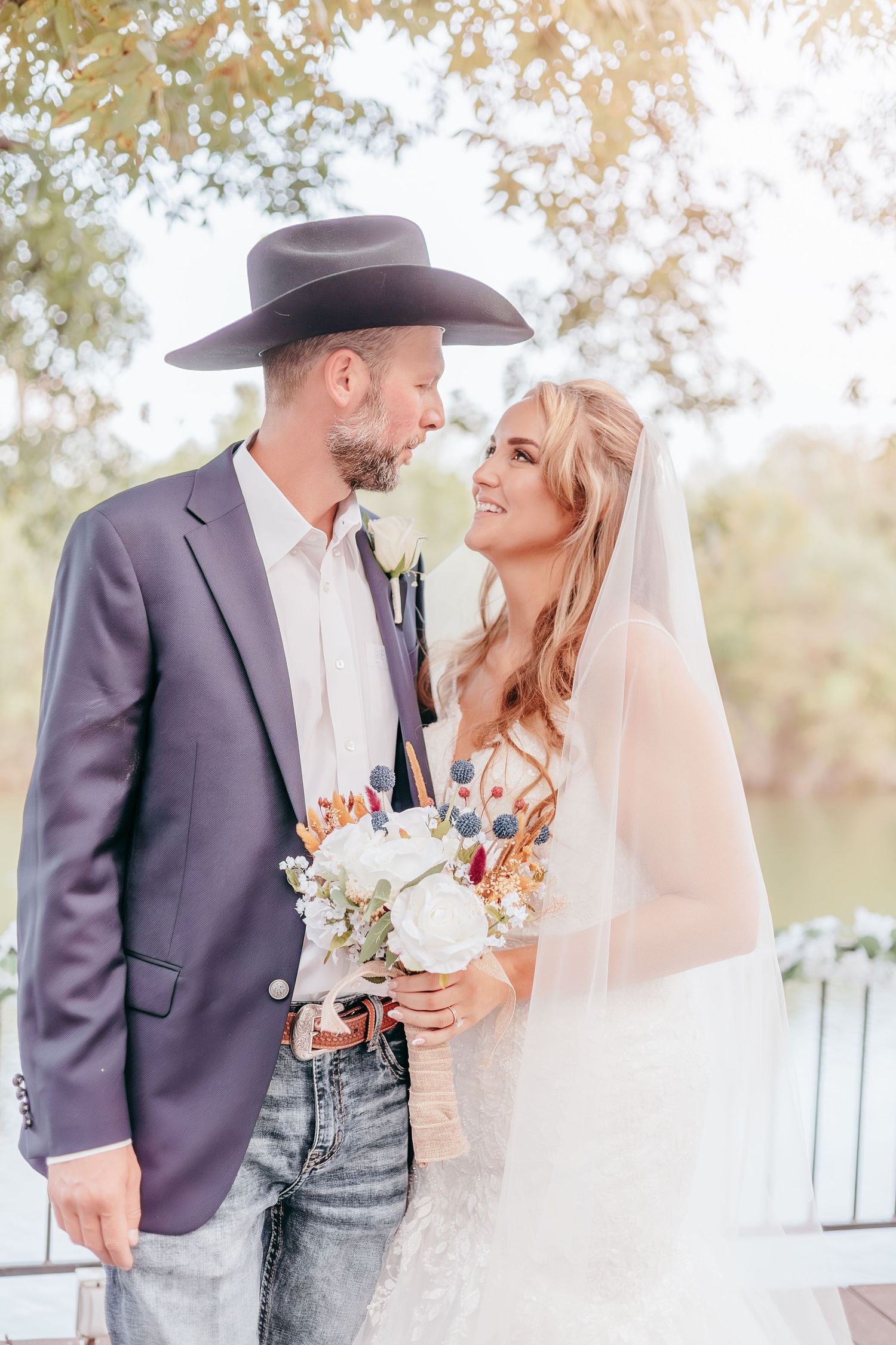 Bride and groom sharing romantic moment outdoors, groom wearing cowboy hat and jeans, bride holding bouquet.