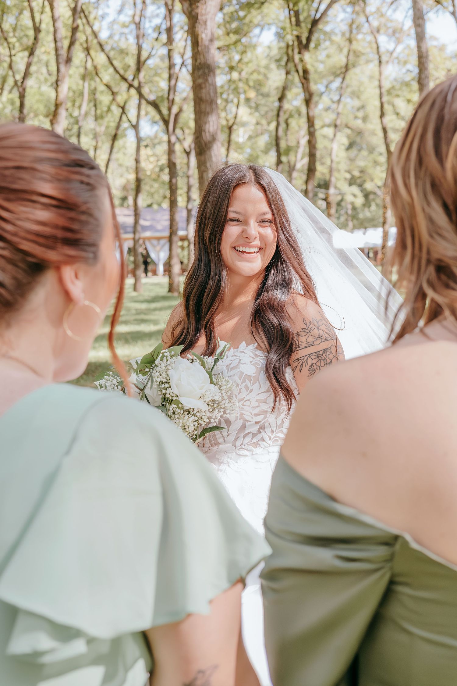 A smiling bride in a lace gown and veil holds a bouquet while speaking with bridesmaids in sage green.