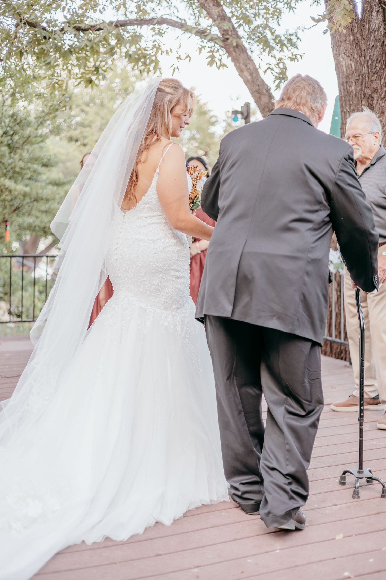Bride in white lace gown and veil walks with groom during outdoor wedding ceremony on wooden deck.