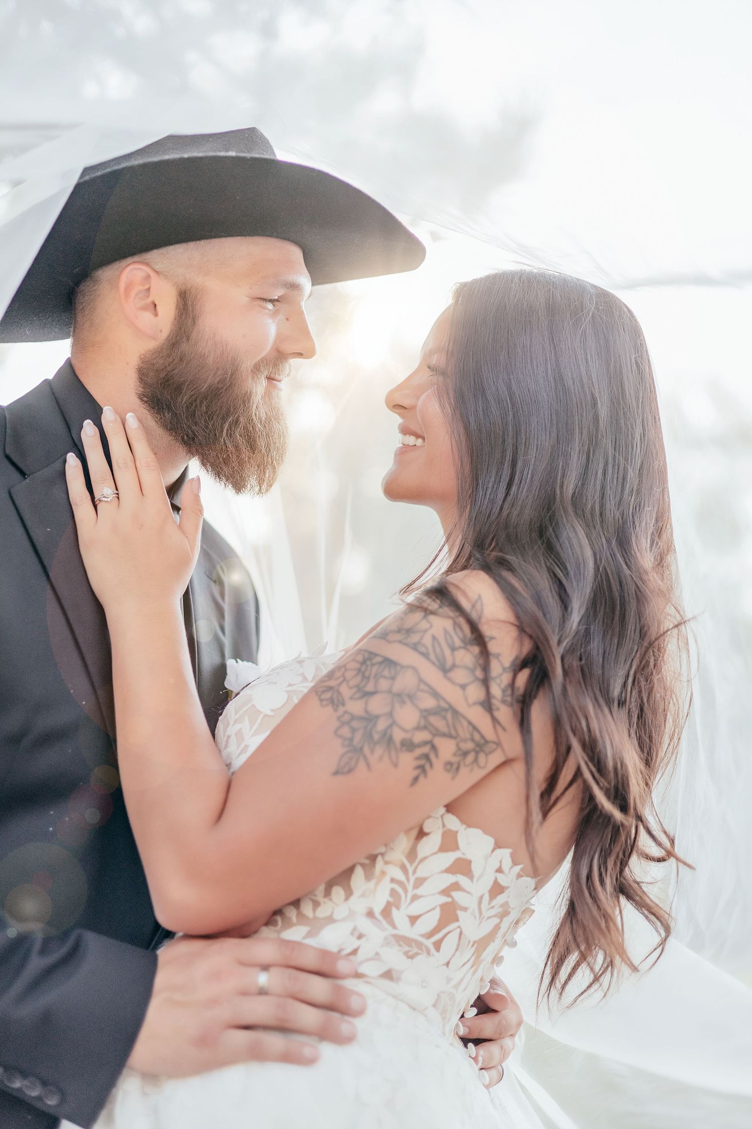 Bride with floral arm tattoo and bearded groom in black cowboy hat smiling at each other under veil.