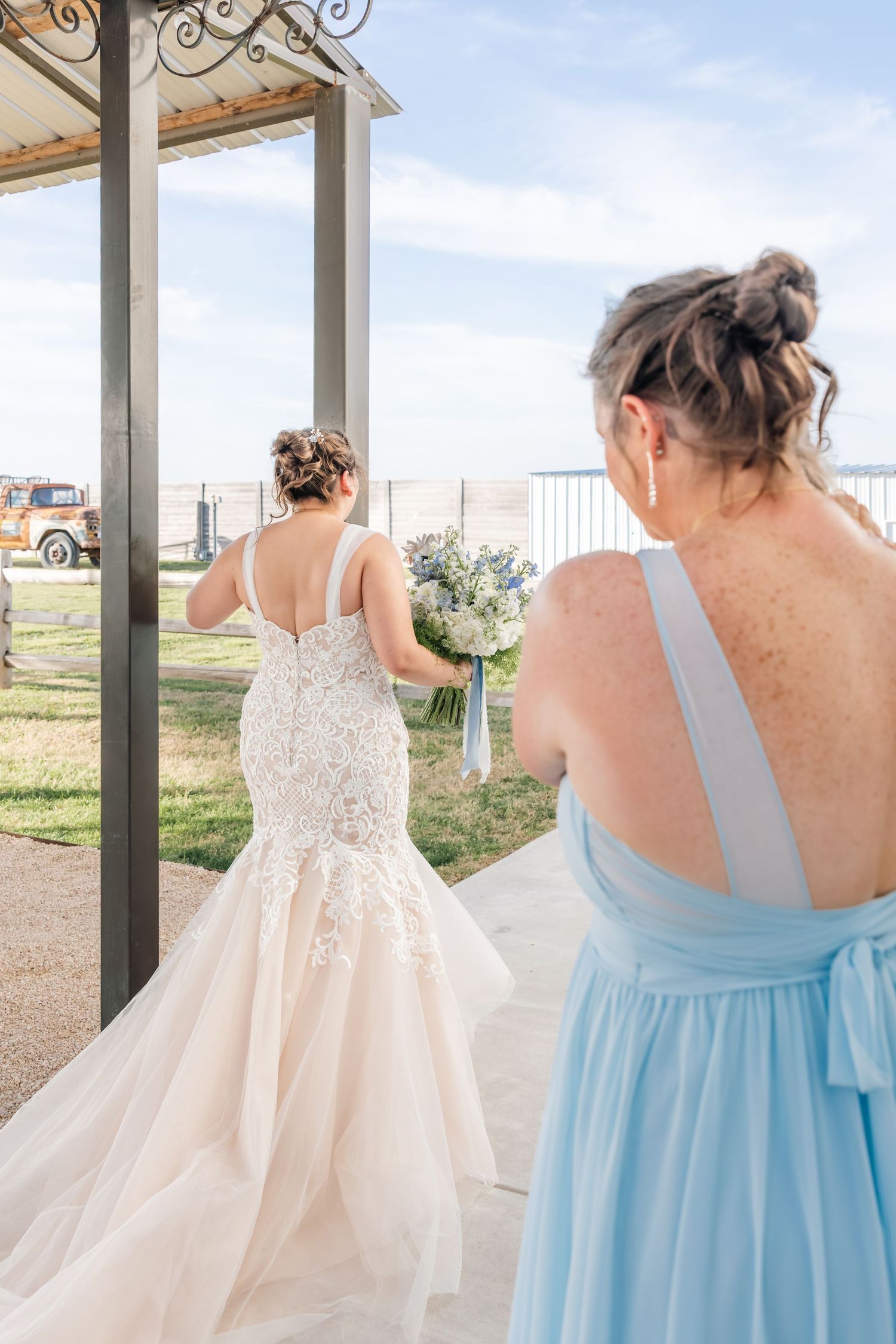 Bride in lace mermaid gown holds blue bouquet while bridesmaid in light blue dress looks on at outdoor wedding venue.