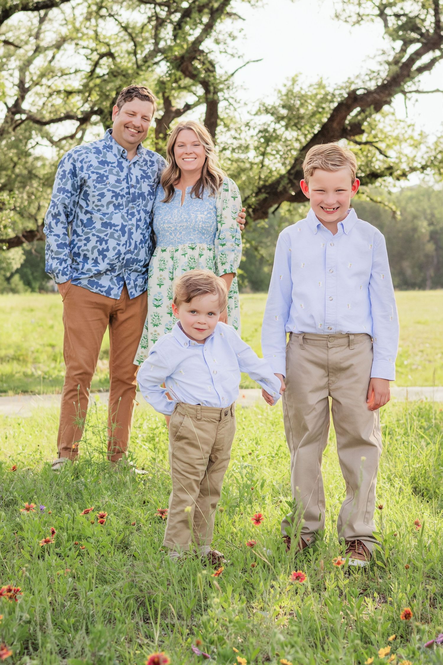 Family of four poses in a wildflower meadow under oak trees, parents and two boys in coordinating blue and khaki outfits.
