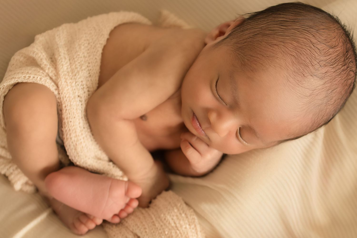 Peaceful newborn baby sleeping curled up on a soft beige blanket, wrapped in a knit wrap.