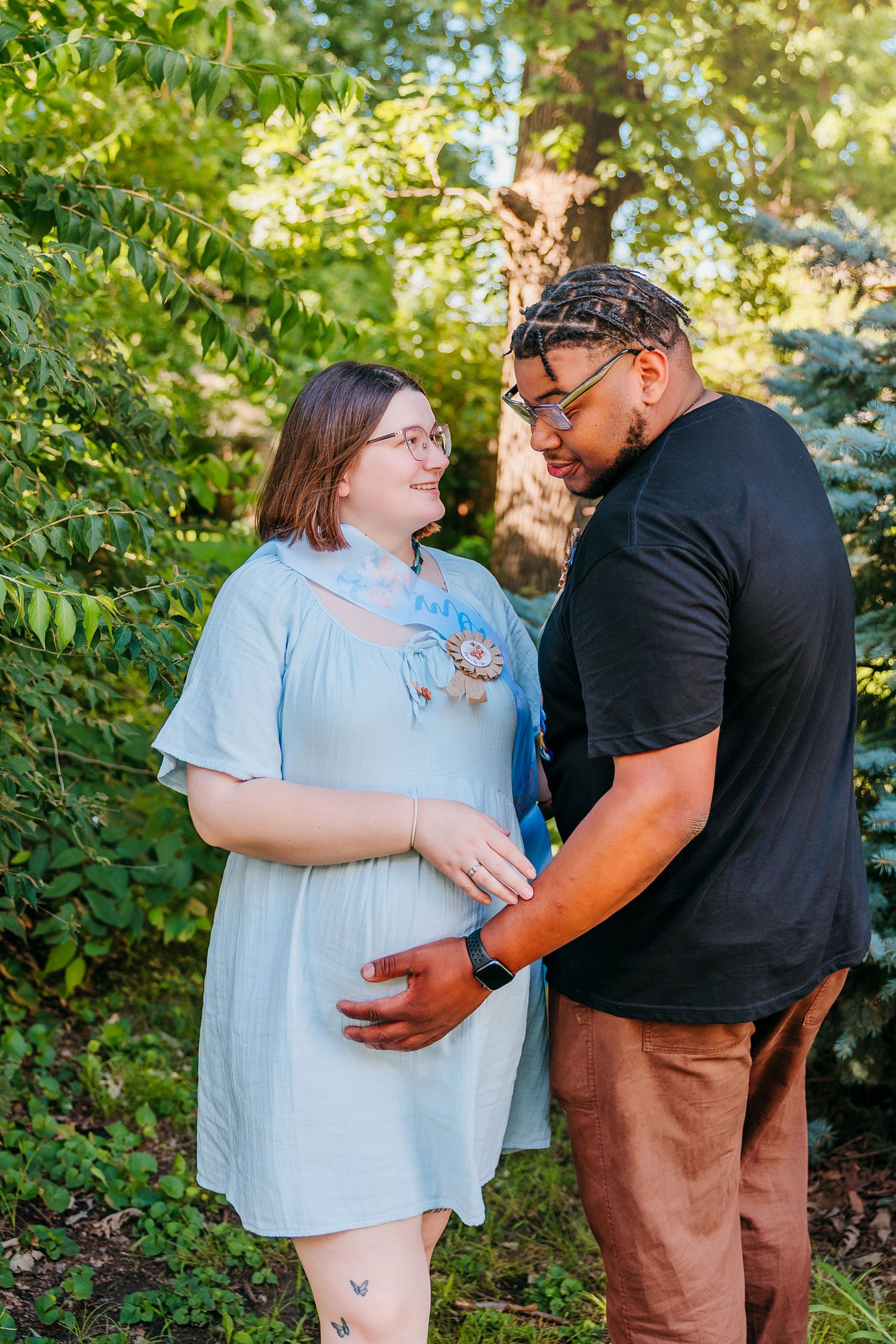Couple sharing a tender moment outdoors, man gently touching pregnant woman's belly amid lush green trees.