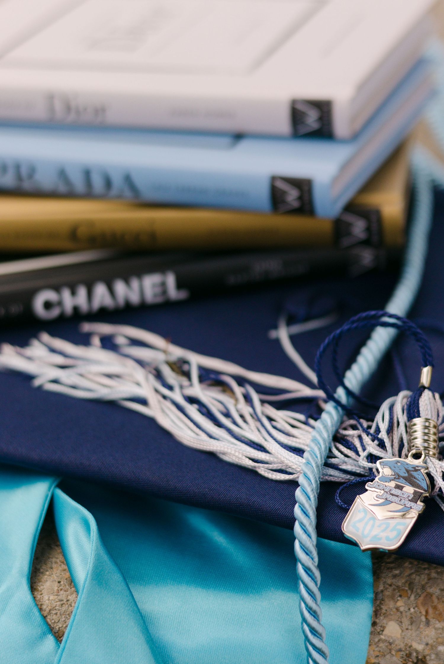 Stack of luxury fashion books with blue graduation cap, cords, and teal sash.