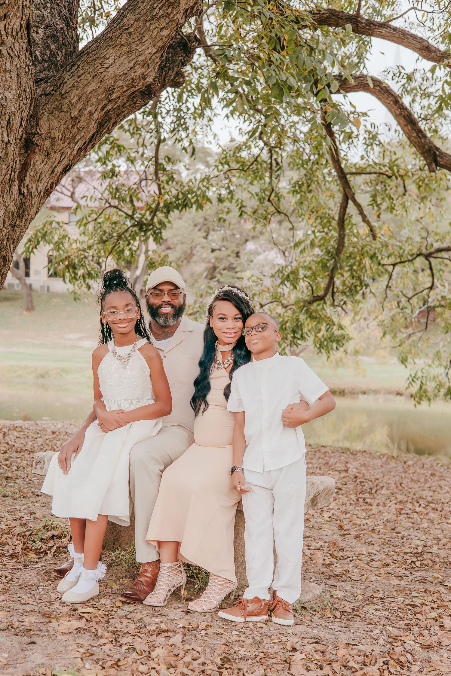 A family of four poses under a tree in coordinated neutral outfits, smiling together on a fall day outdoors.