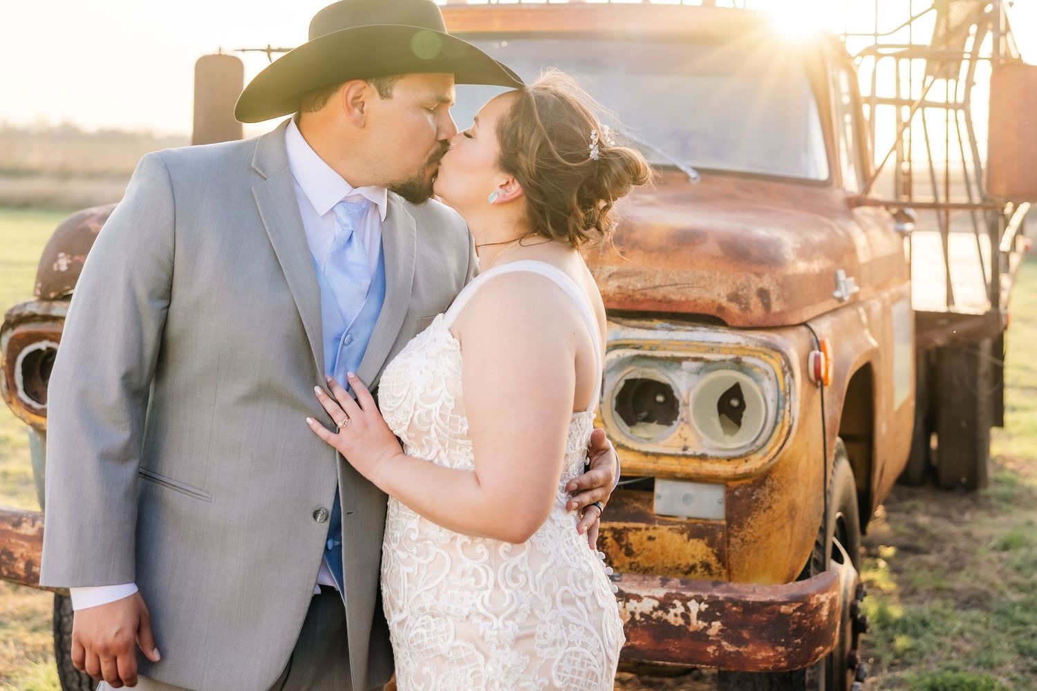 Groom in cowboy hat and bride share a kiss next to a weathered vintage truck at sunset.