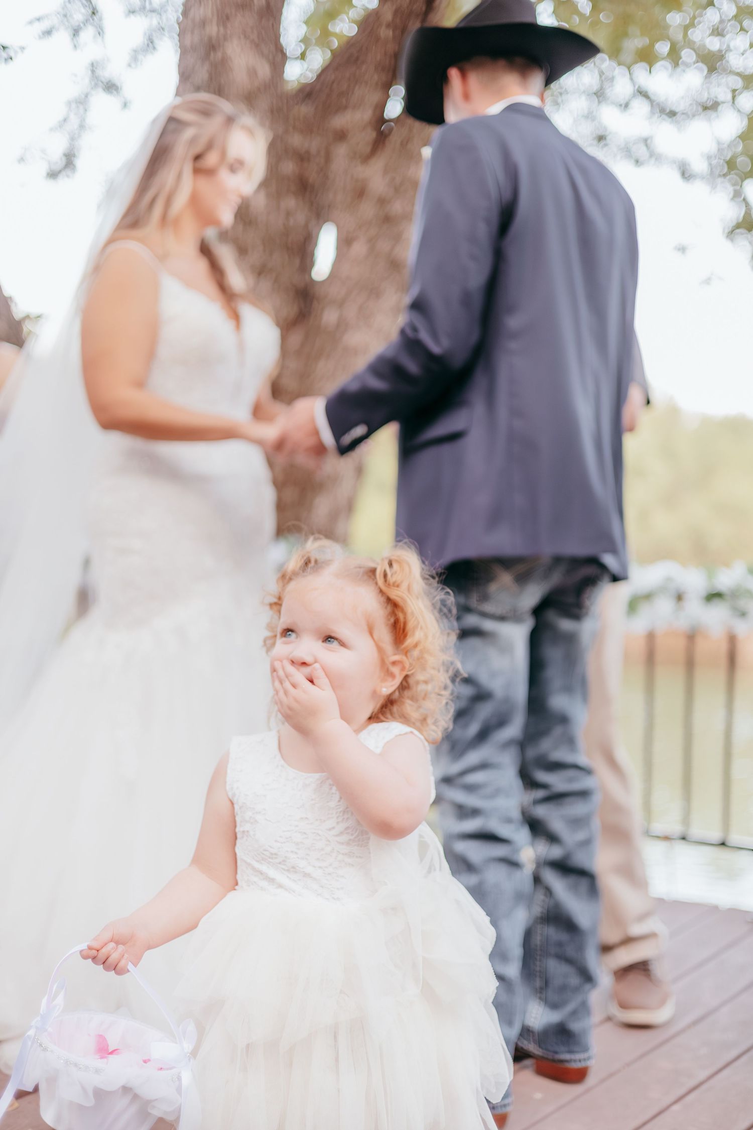Curly-haired flower girl in white dress at outdoor wedding ceremony with bride and groom holding hands in background.