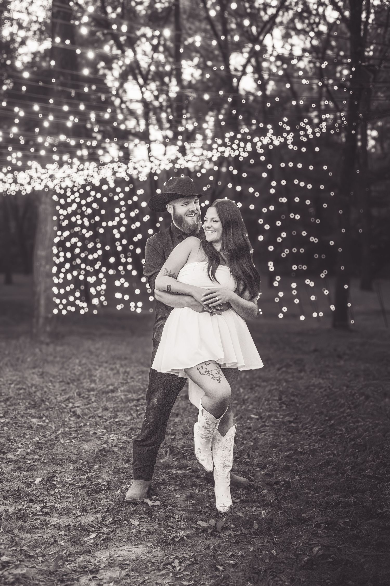 Black and white romantic photo of a couple dancing and laughing before a twinkling lights backdrop.