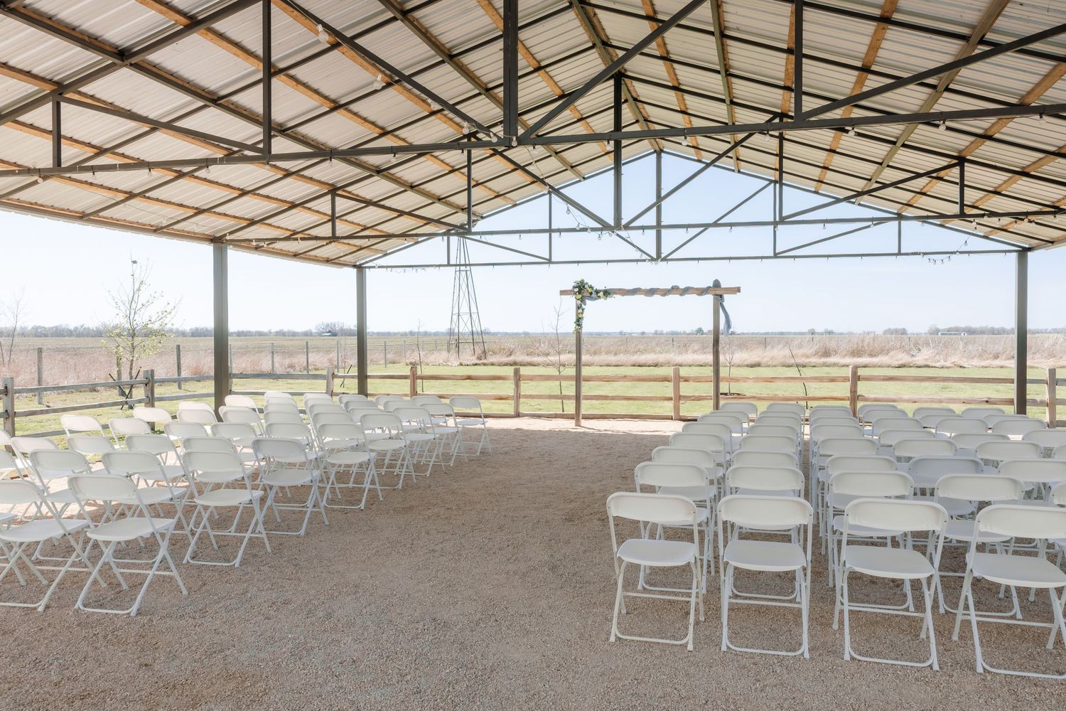 Rustic outdoor wedding ceremony setup with white folding chairs under an open-air barn structure.