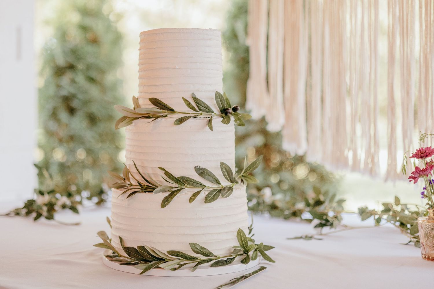 Elegant three-tier white wedding cake adorned with olive branch greenery on a table with floral decorations.