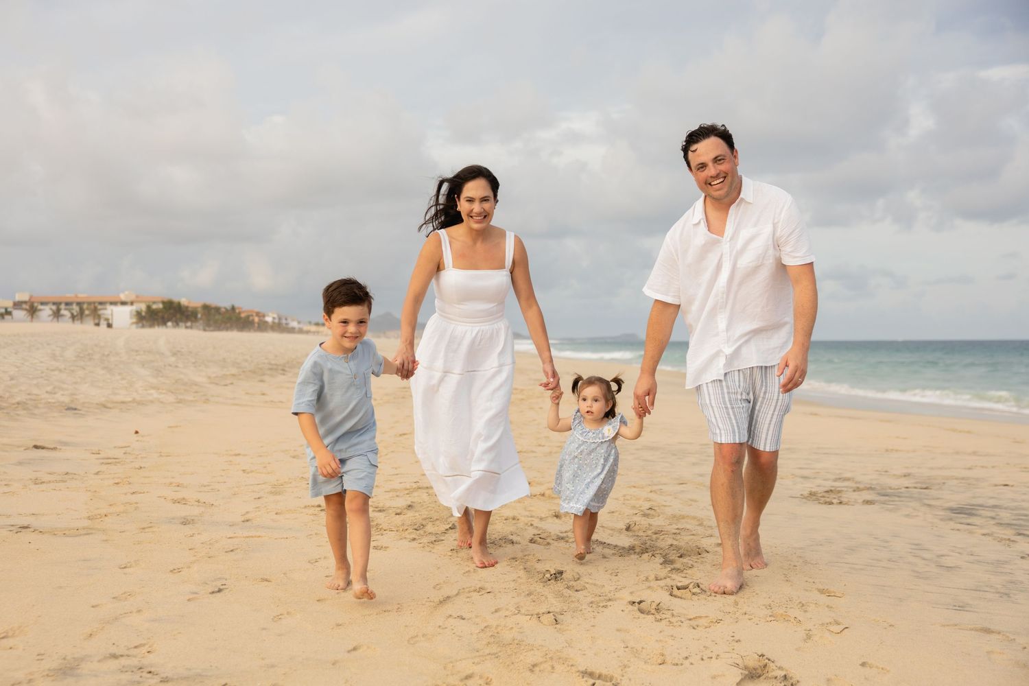 Happy family of four walking barefoot on a sunny sandy beach, parents holding hands with two young children.