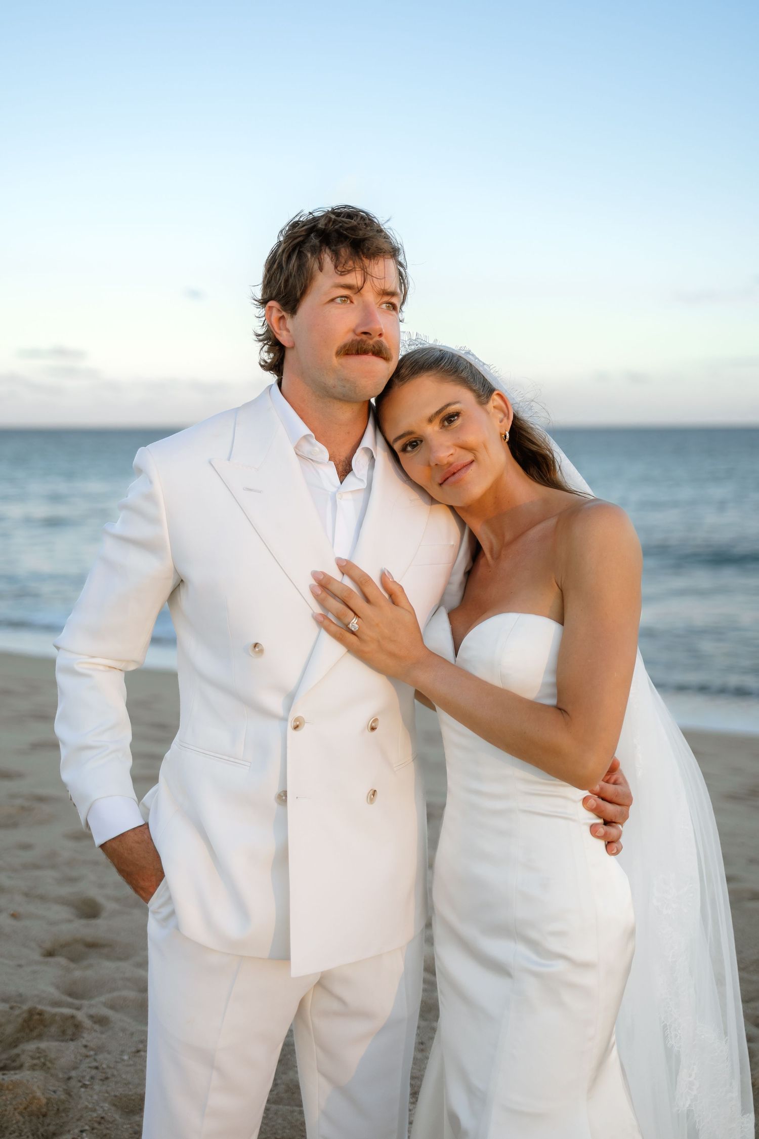 Wedding couple in white embracing on sandy beach at sunset.