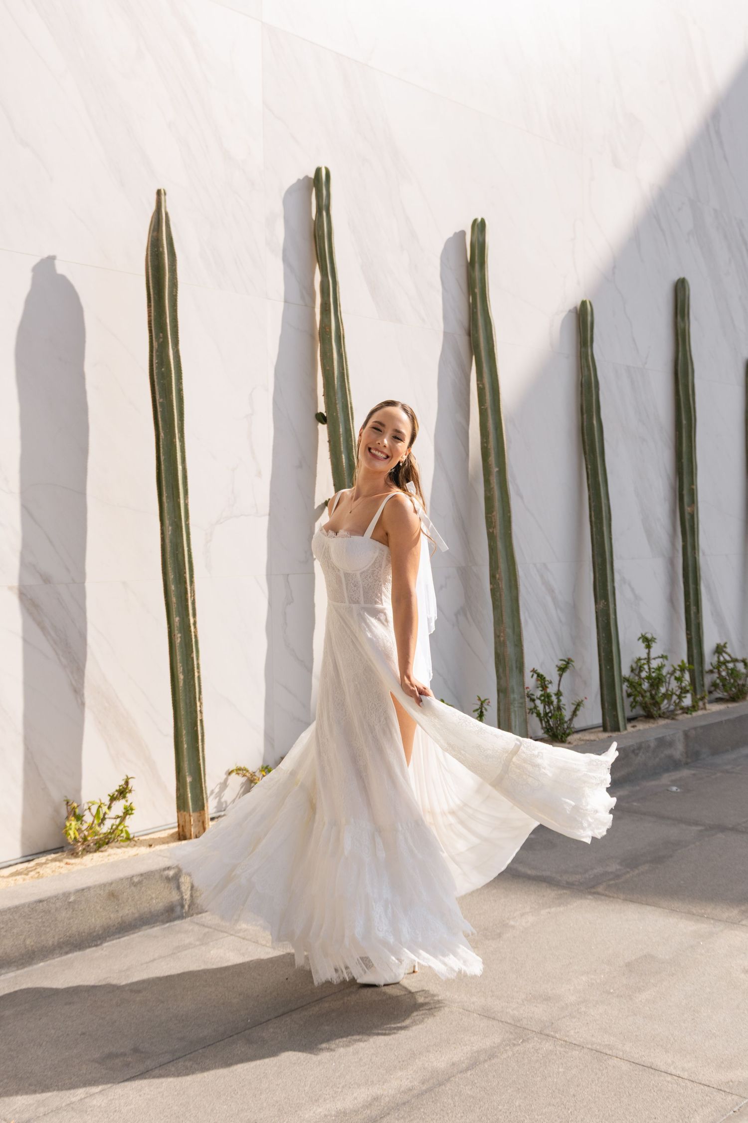 Bride in flowing white gown poses against white wall with tall green plants.