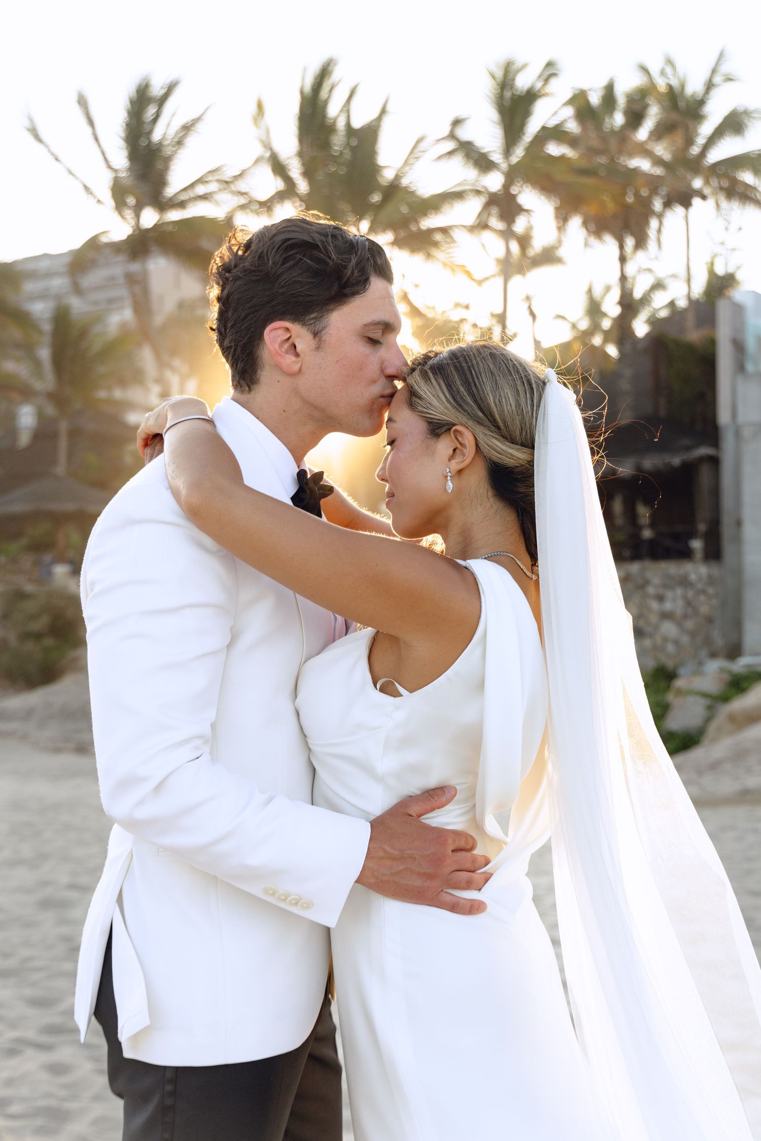 Newlywed couple sharing romantic kisses on a tropical beach with palm trees at sunset.