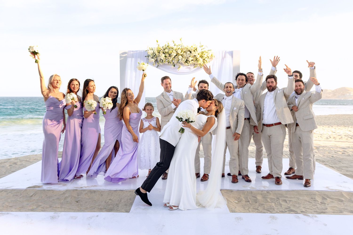 Joyful wedding party celebrating on beach with arch and ocean backdrop.