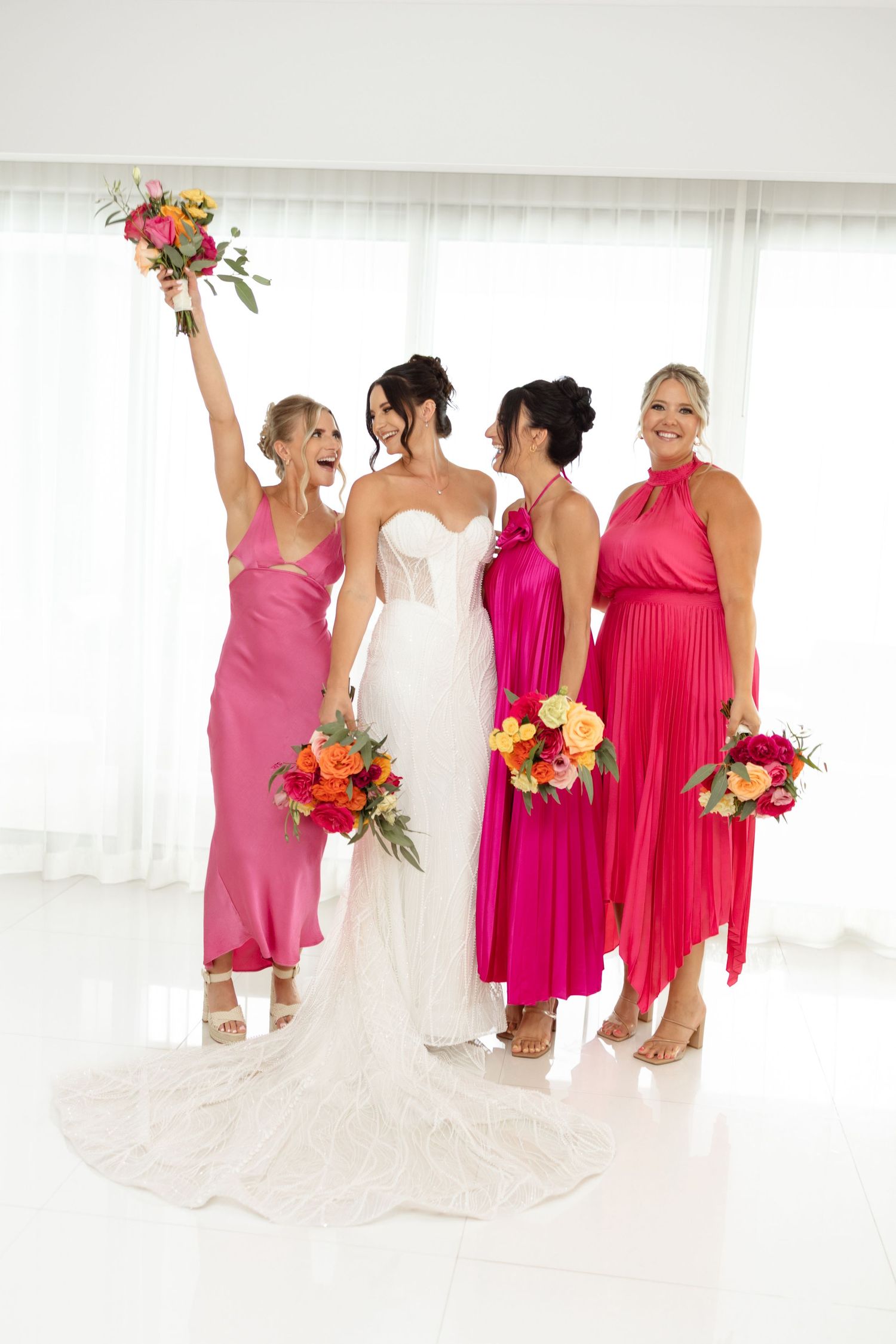 Wedding party in pink dresses posing with colorful bouquets against white backdrop.