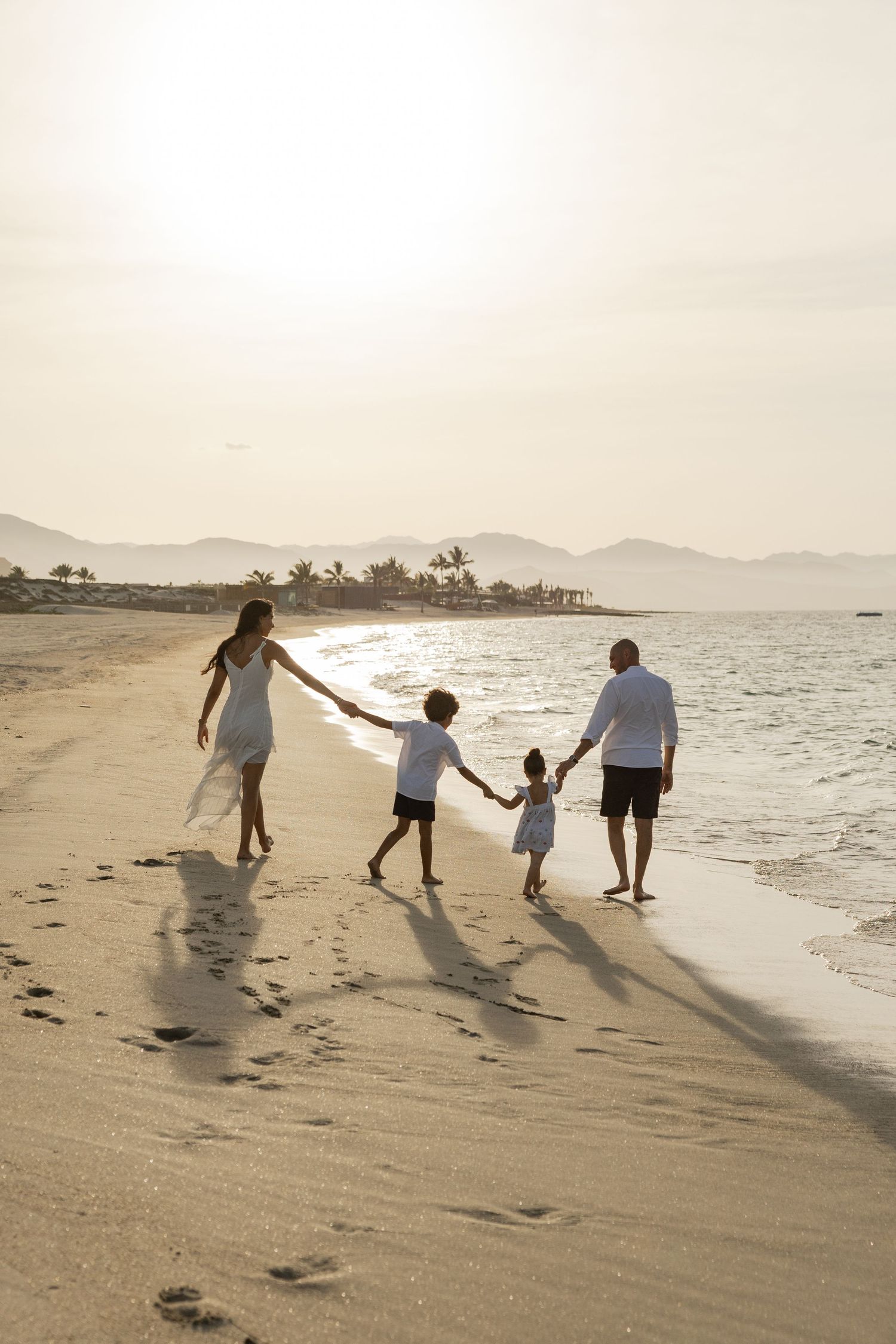 Family of four holding hands walking along a sandy beach at sunset with mountains in the background.