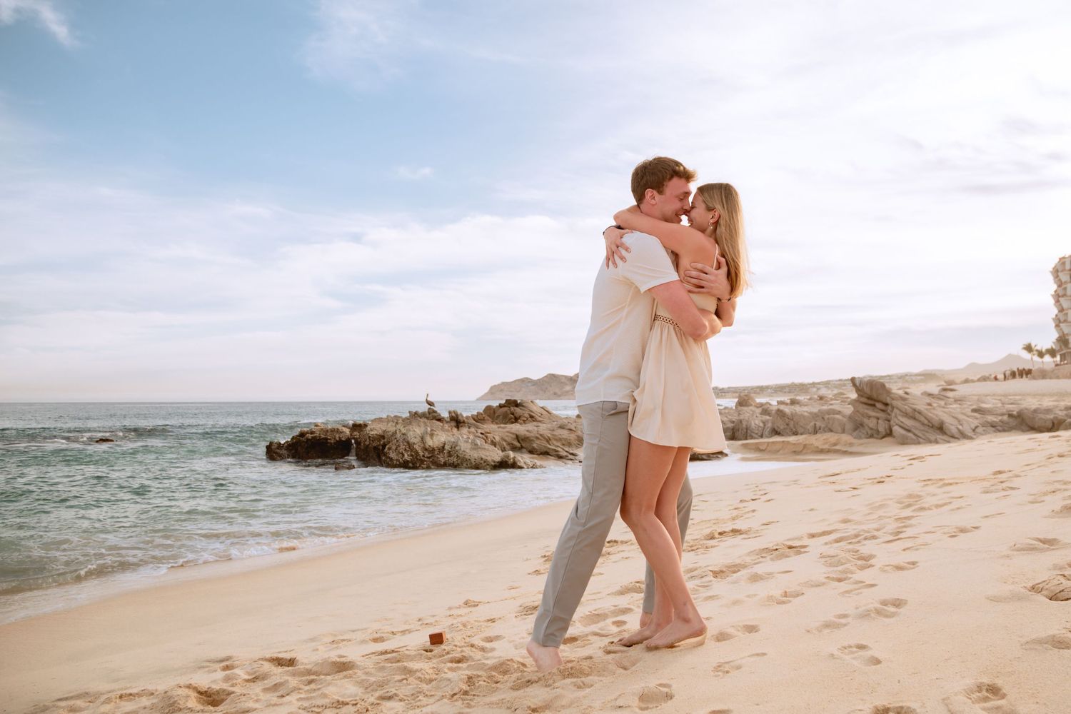 Romantic couple in light colored outfits embrace on a tropical beach with ocean waves and rocks in the background.