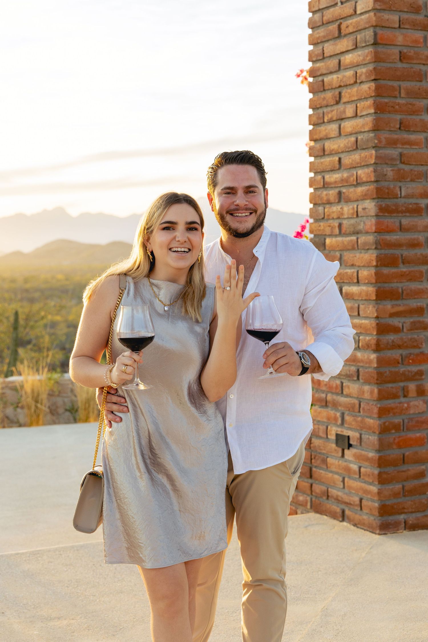 A couple shares wine and laughter while enjoying sunset views from a brick terrace overlooking scenic hills.