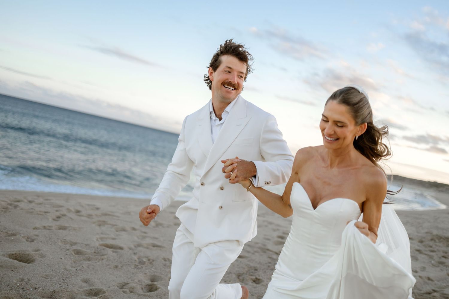 Couple in white attire running playfully along a beach at sunset during their wedding celebration.