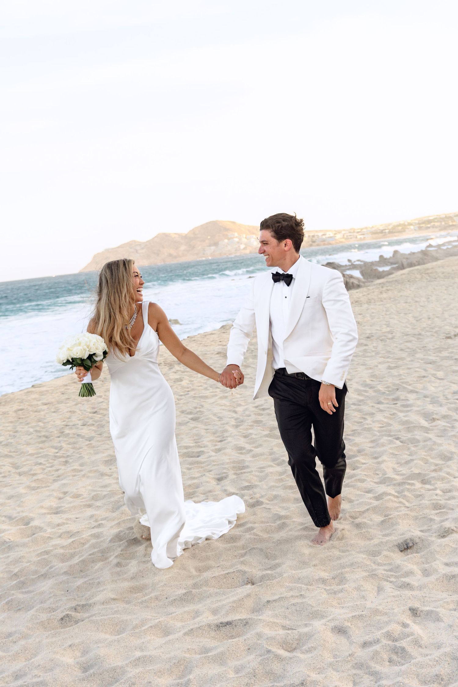 Wedding couple walking barefoot on sandy beach holding hands with mountains in background.