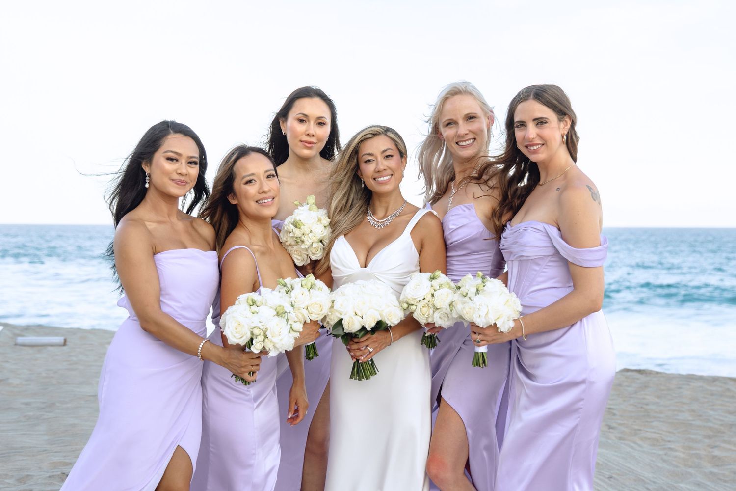 Bridesmaids in lavender dresses posing together on beach with bouquets.