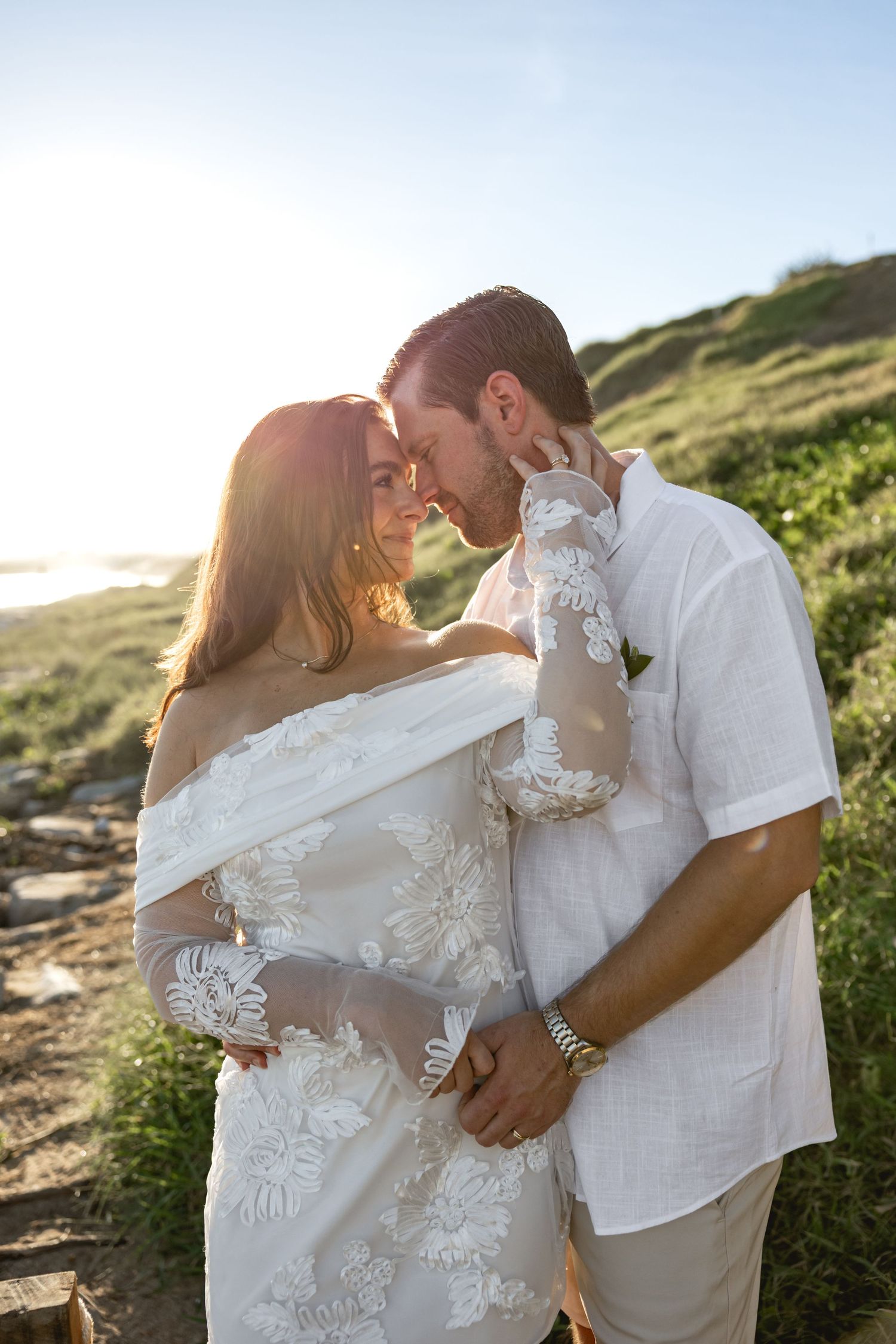 Couple in wedding attire sharing a romantic moment at sunset near a coastal hillside, bride in lace gown.