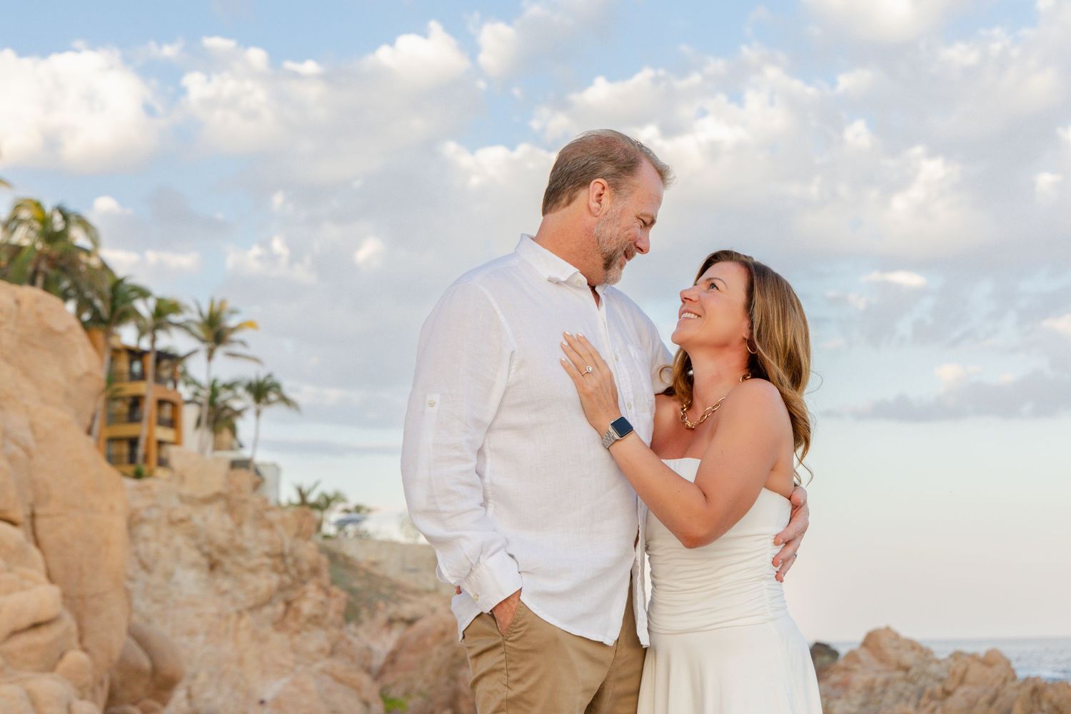A couple shares a romantic moment on a beach at sunset with palm trees and ocean views in the background.