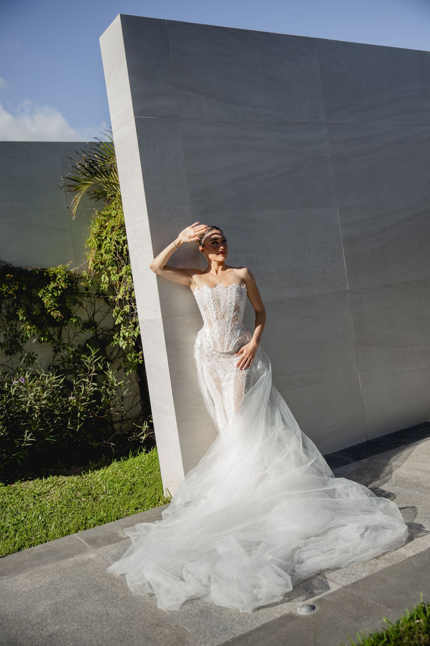 Bride in elegant white tulle mermaid wedding gown with lace corset bodice posing against modern concrete wall outdoors.